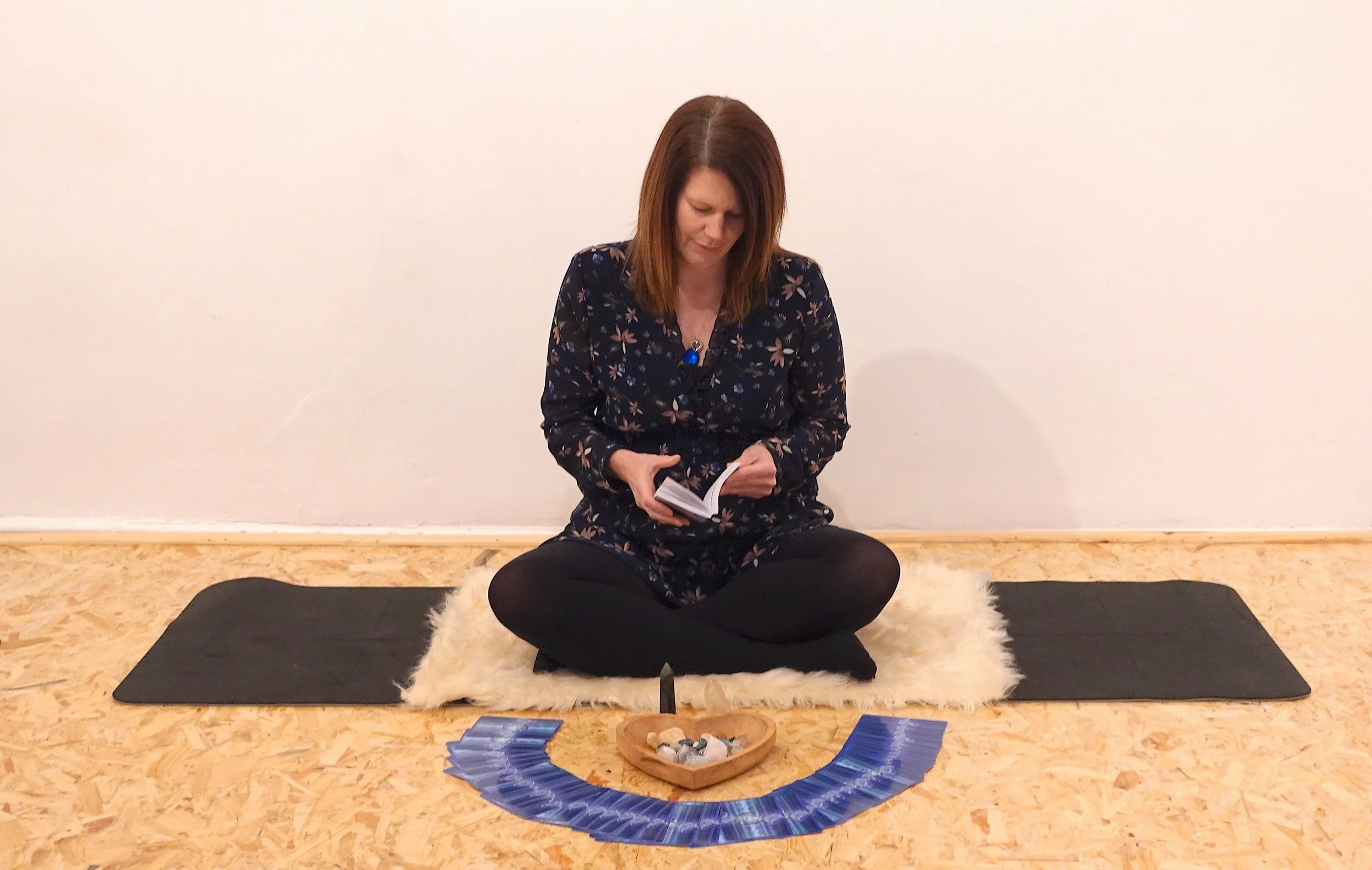 Woman sitting cross-legged on a white furry rug with tarot cards, crystals, and a knife on a wooden floor against a plain white wall.