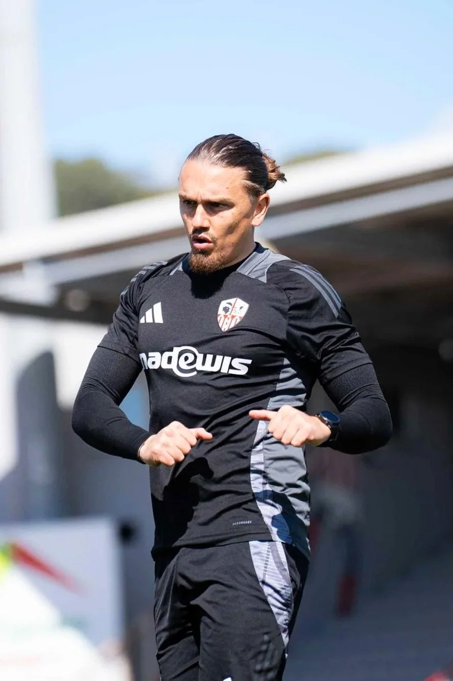 un homme avec les cheveux longs et une barbe, portant un maillot de sport noir avec le logo du club de football Sevilla FC, en train de courir ou faire de l'exercice en plein air