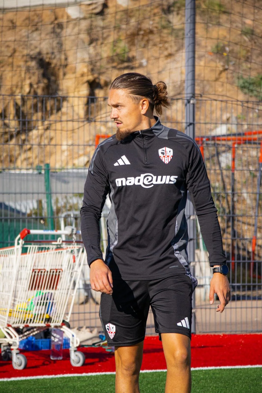 Un homme portant un survêtement de sport noir avec des logos Adidas et l'emblème du club de football souligne, se tenant sur un terrain d'entraînement. Arrière-plan avec une clôture métallique, un panier de course et un mur rocheux.