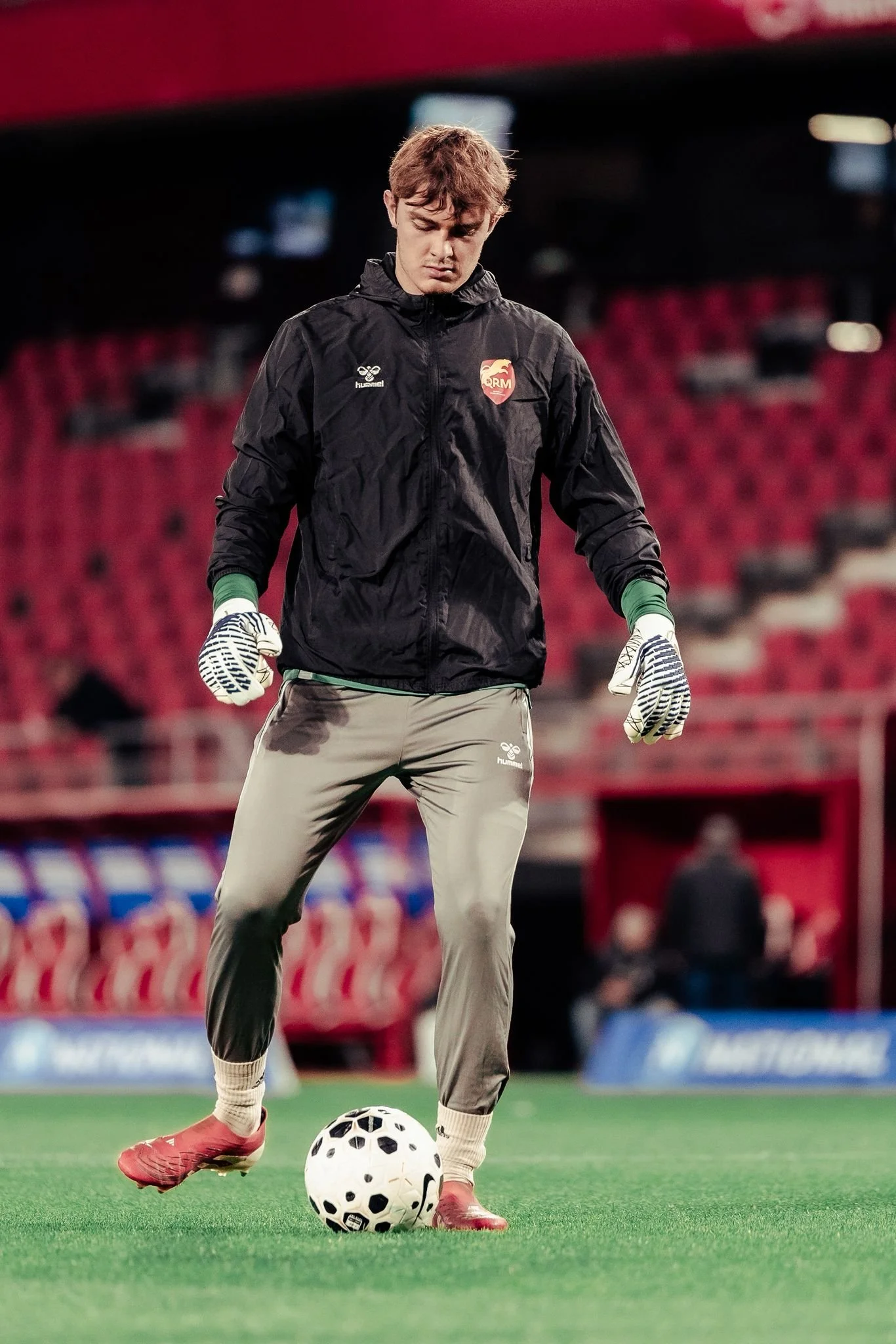 Un gardien de but en tenue d'entraînement, portant des gants, une veste noire et un pantalon gris, se tient sur un terrain de football avec un ballon à ses pieds dans un stade vide.