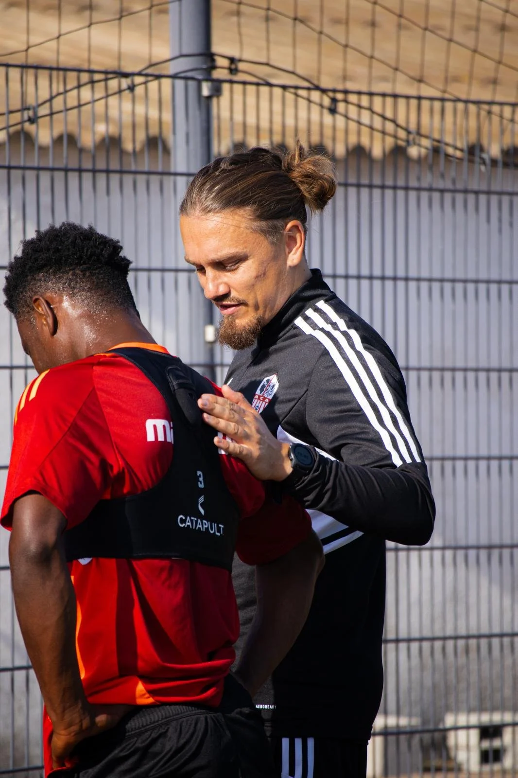 Un entraîneur donne des instructions à un joueur de football lors d'une séance d'entraînement, dans une enceinte grillagée.