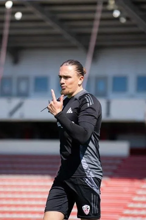 Un homme en tenue de sport noir sur un terrain d'athlétisme, faisant un signe de la main, avec un bâtiment en arrière-plan.