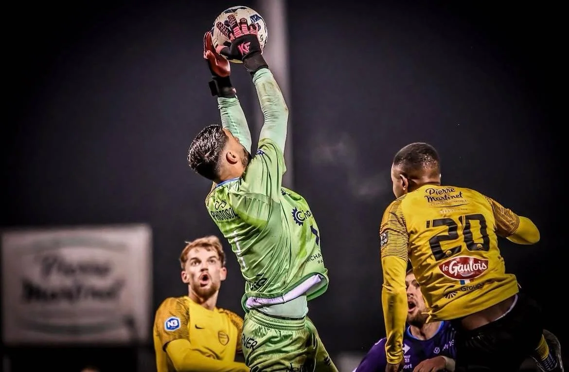 Un gardien de but en vert attrape un ballon lors d'un match de football. Deux joueurs en jaune et un autre en violet observent l'action, sous un éclairage artificiel intense.