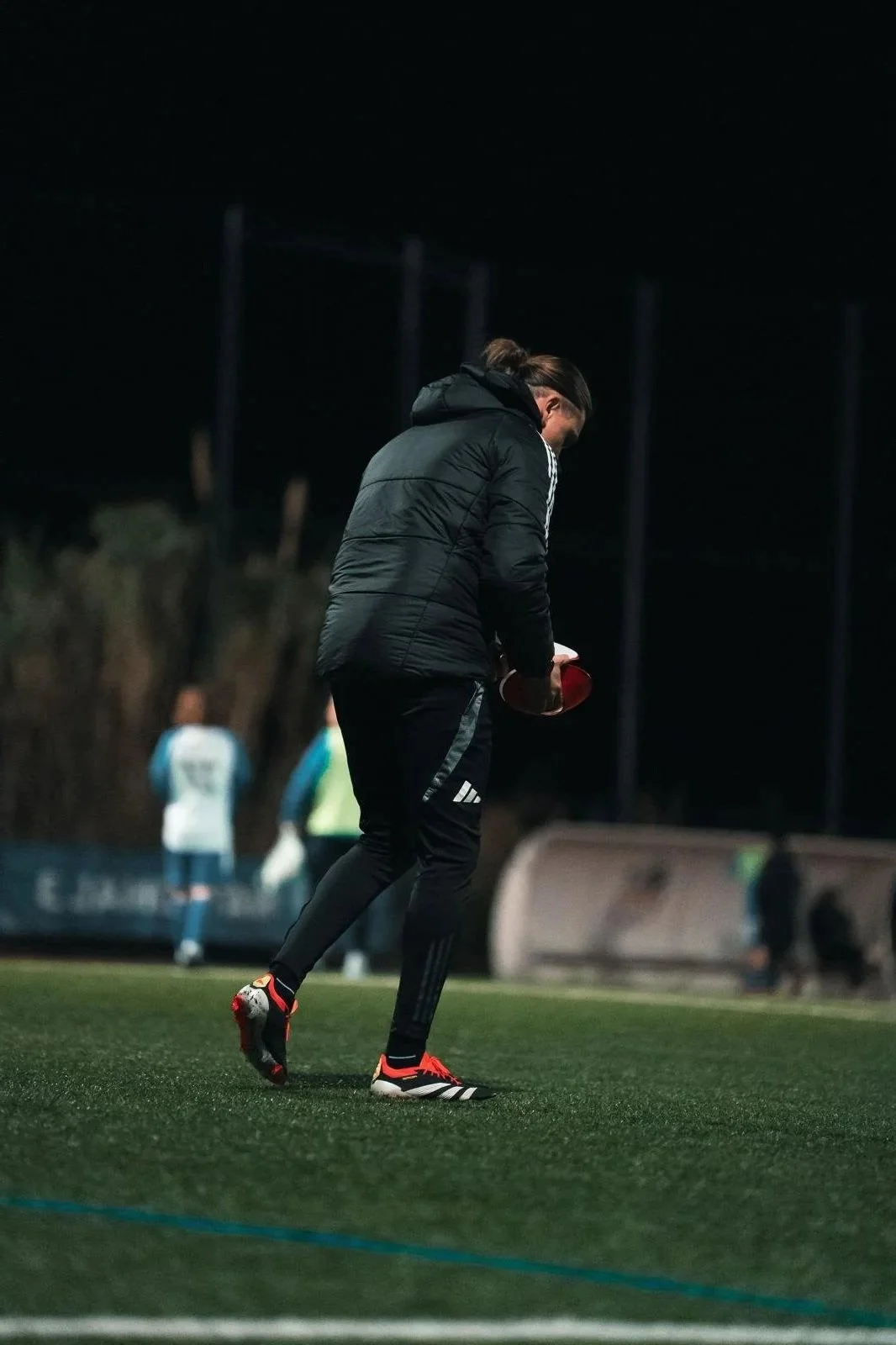 Une personne habillée en équipement de football, tenant un trophée, sur un terrain de football la nuit, avec d'autres personnes en arrière-plan.