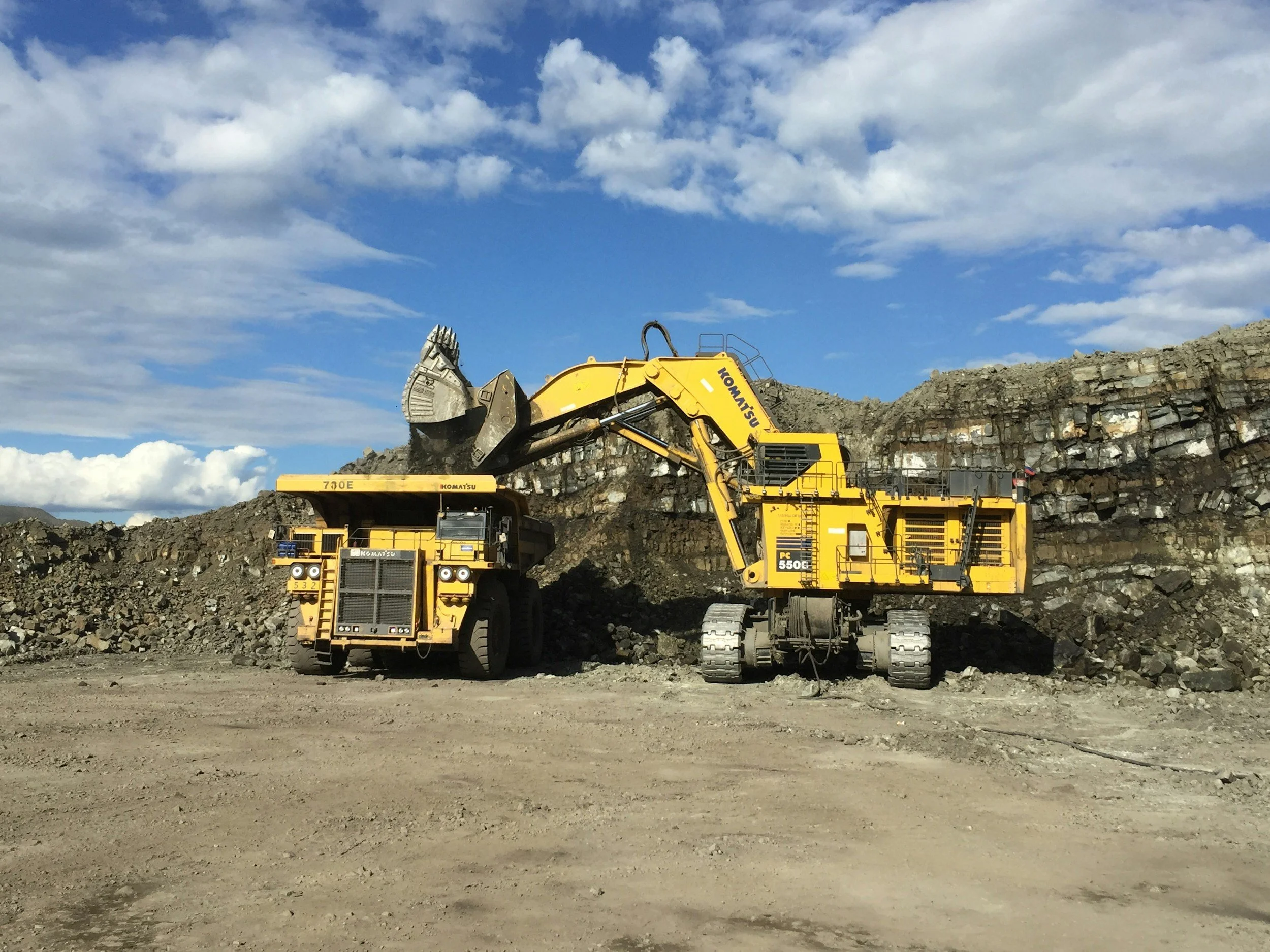 A large yellow excavator moving rocks into a dump truck at a mining site under a partly cloudy sky.