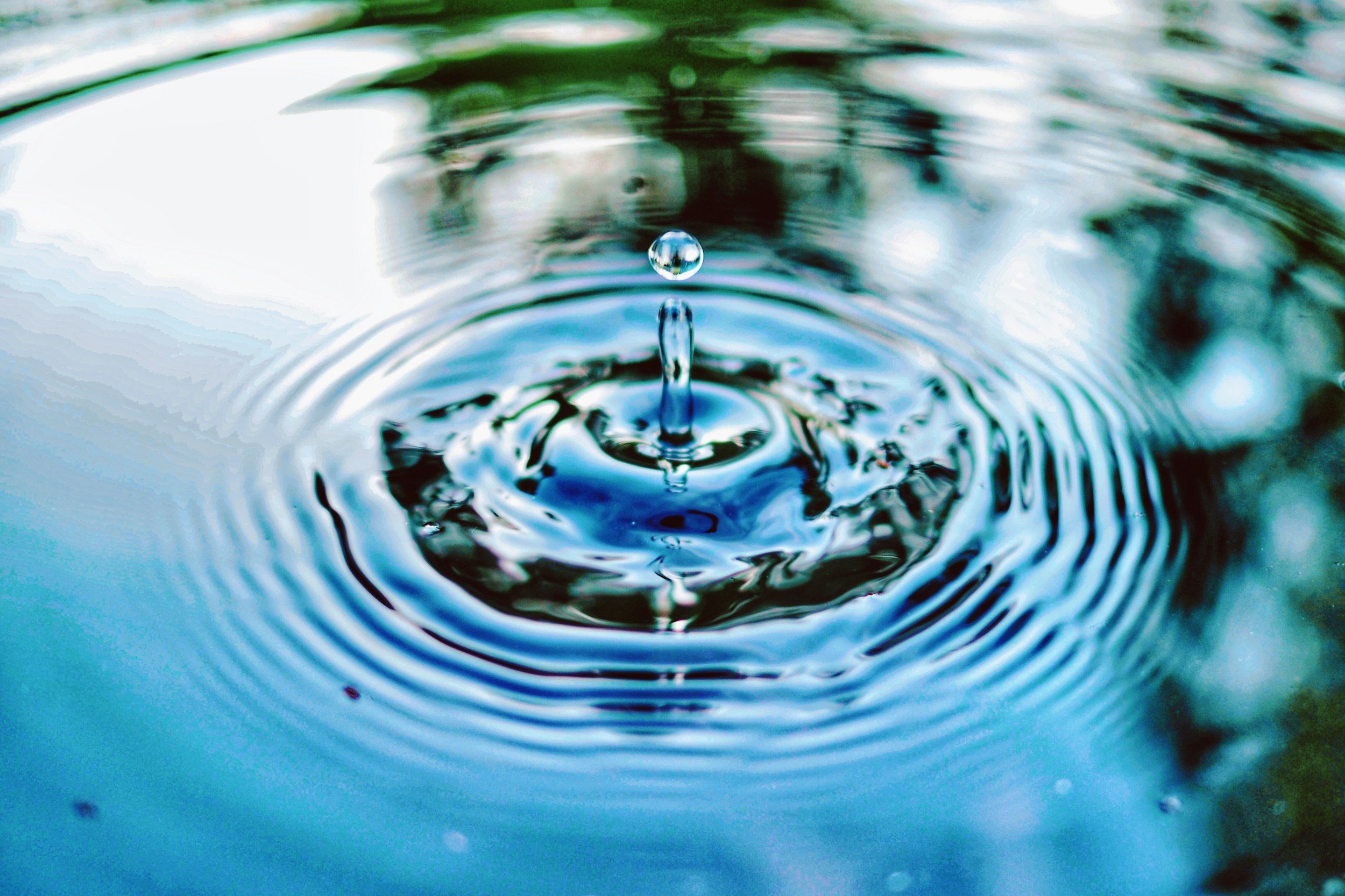 A close-up of a water droplet falling into a body of water, creating ripples and a splash, with reflections of the surroundings.