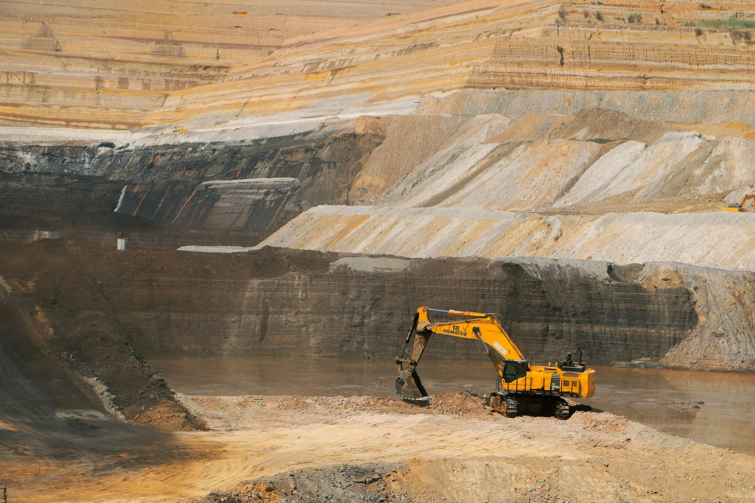 A yellow excavator working in a large open-pit quarry, with multi-colored layers of rock and earth in the background.
