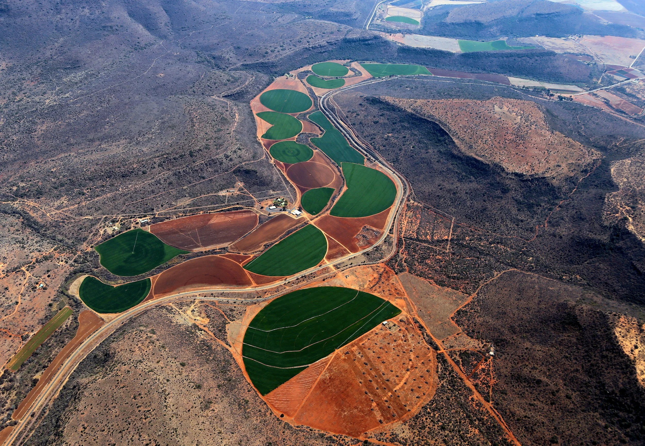 Aerial view of farmland in a semi-arid landscape with green crop fields surrounded by brown, barren land and sparse shrubbery.