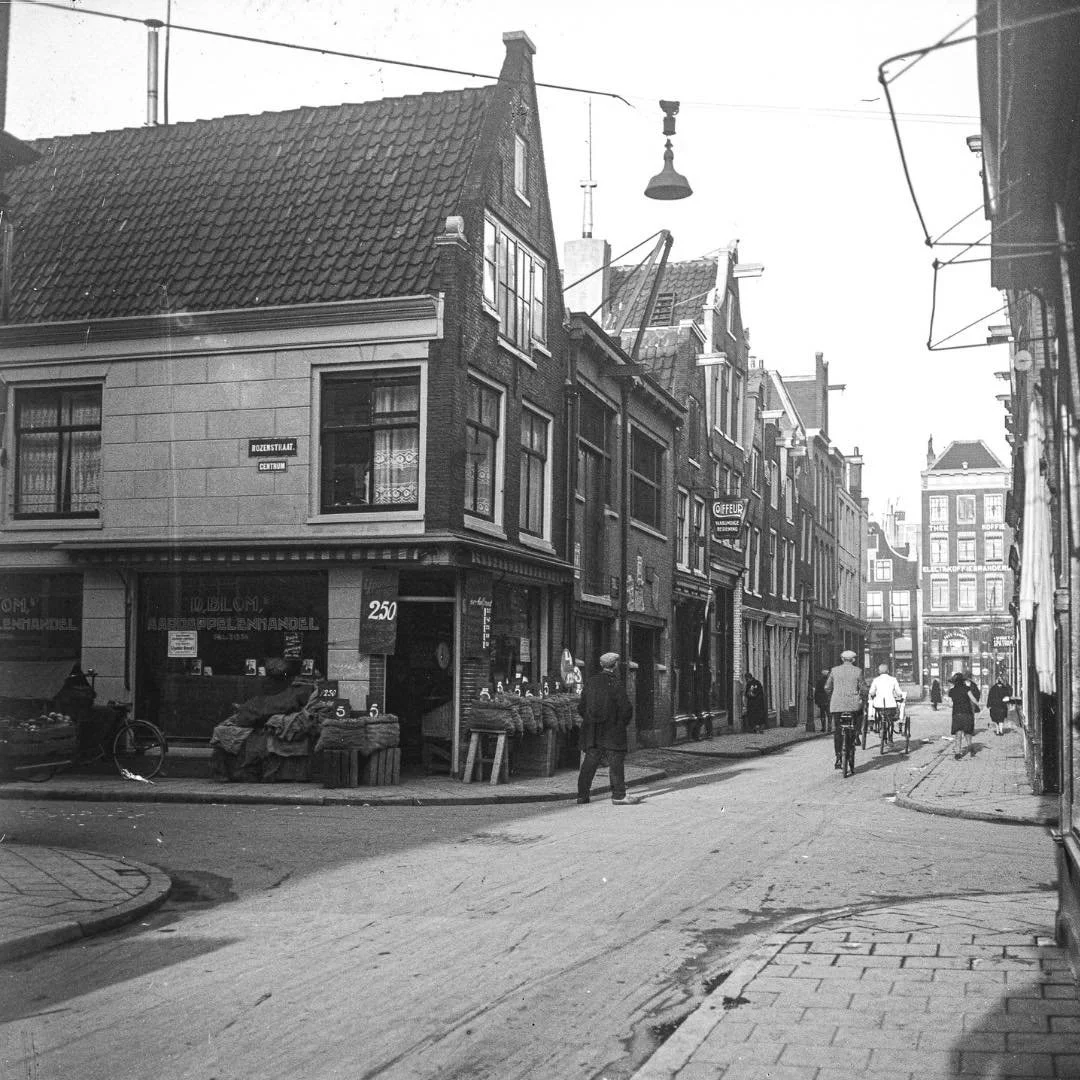 1929 &amp; Now: let's travel 96 years back in time and see what the Jordaan used to look like back in the day. In this specific photo you see Eerste Rozendwarsstraat on the corner with Rozenstraat. The corner building at Eerste Rozendwarsstraat 22 wa