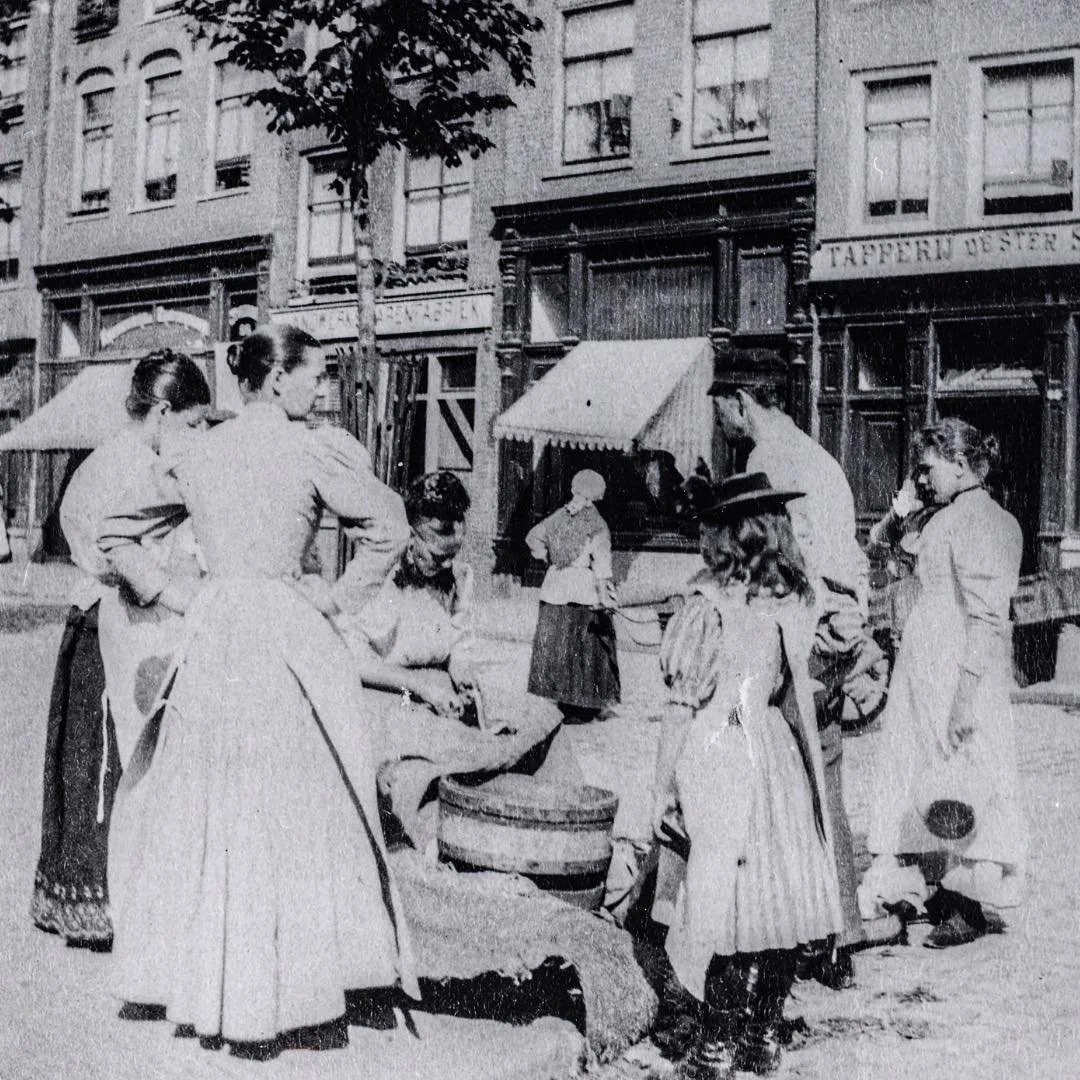 1910 &amp; Now: in this photo you see the houses of Elandsgracht 92-82, close to the entrance of Hazenstraat. In the center of the photo, a fish seller is busy cleaning some fish while a group of ladies gather around her. #thenandnowamsterdam