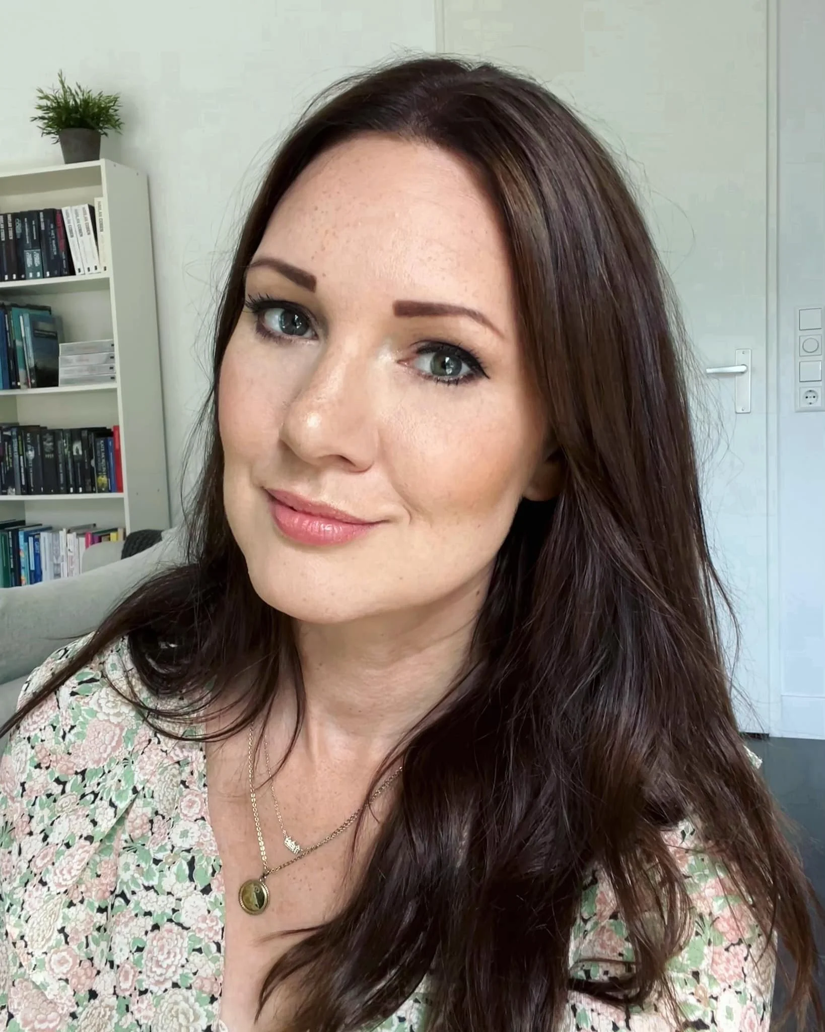 A woman with long, wavy brown hair and blue eyes sitting in a room, wearing a floral top and layered necklaces.