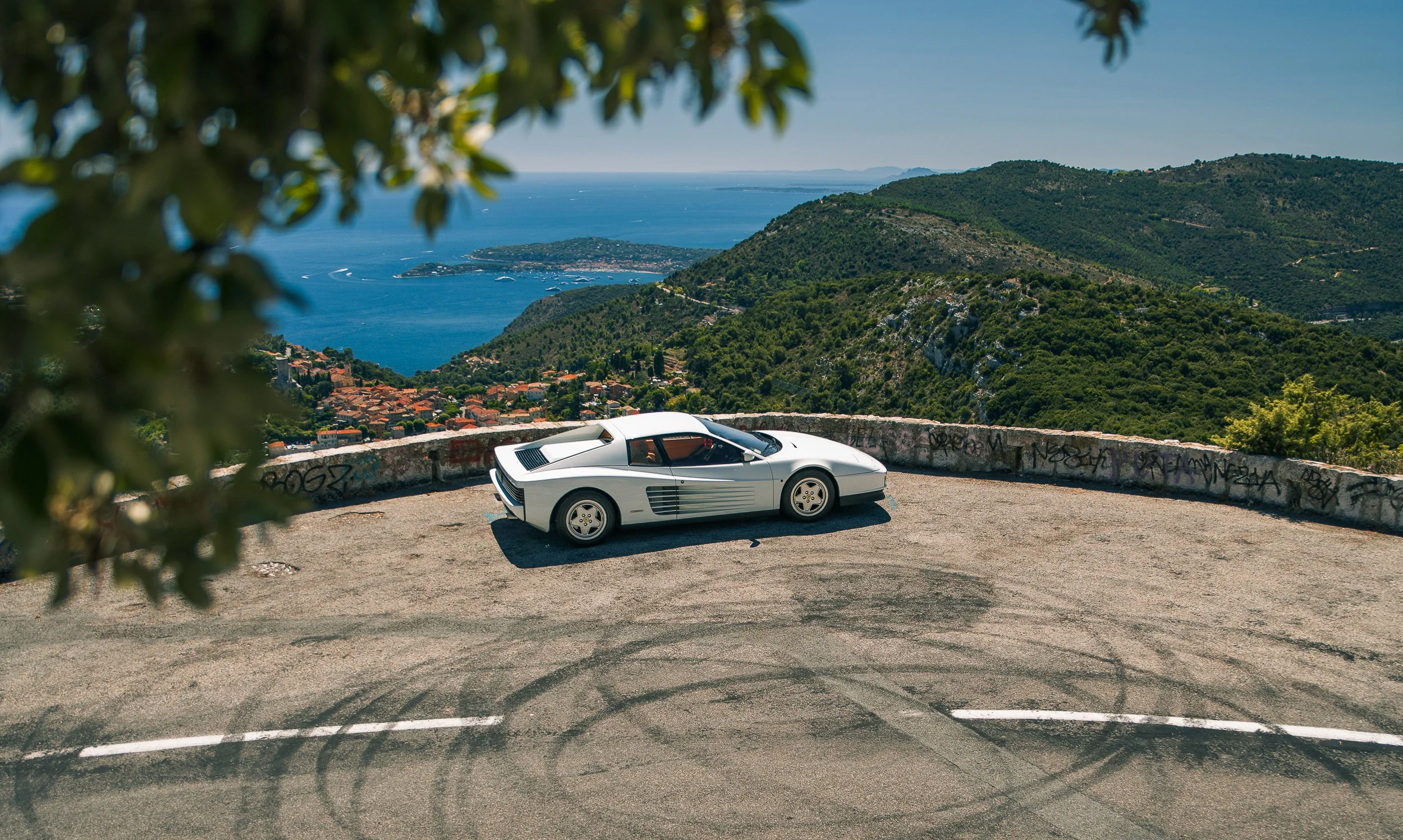 A white vintage sports car parked on a mountain road with a scenic view of the ocean and hills in the background, partially obscured by tree branches.