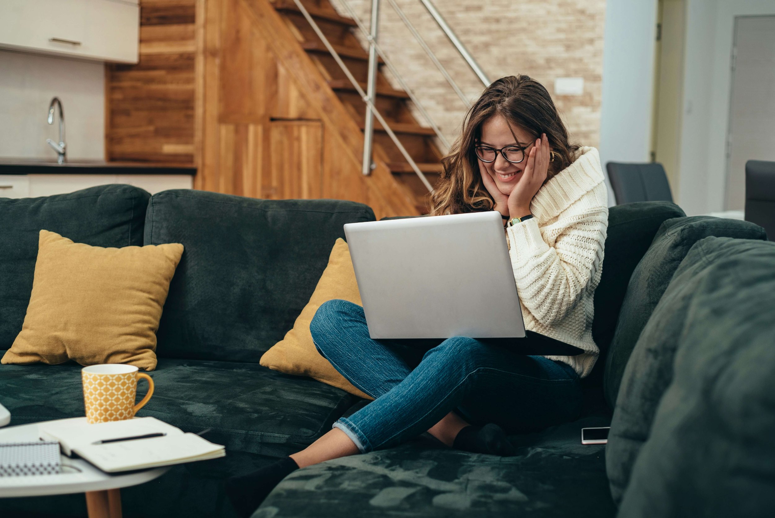 A woman sitting on a dark green couch, smiling with her hands on her face, looking at a laptop. She is wearing glasses, a beige sweater, and blue jeans. There are yellow pillows on the couch, a yellow patterned mug, notebooks, and a smartphone nearby. The background features a wooden staircase and kitchen.
