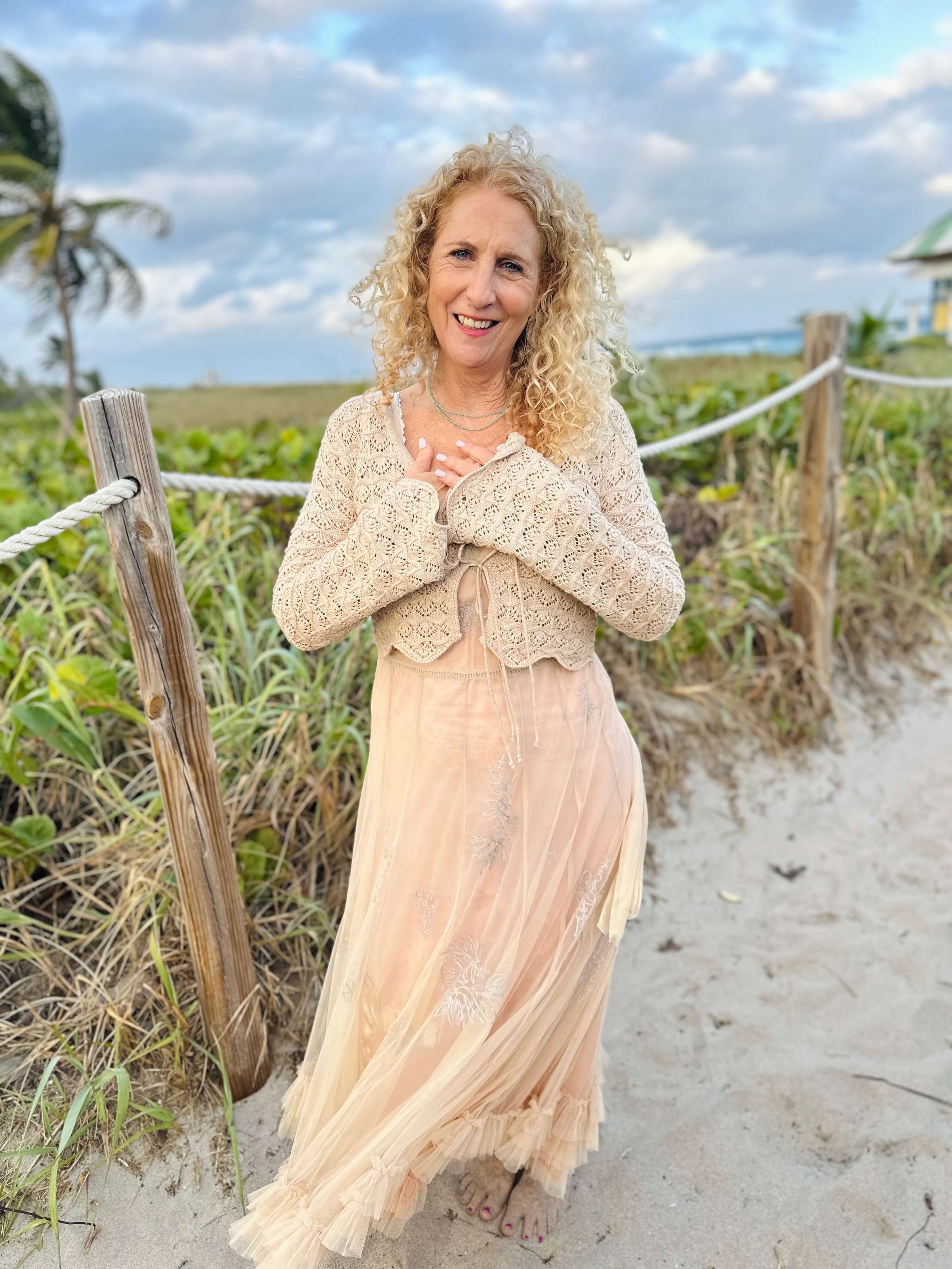 Jozi Radus standing on a sandy beach near a wooden fence with lush greenery, blue sky with clouds, and a house in the background, smiling and looking at the camera.