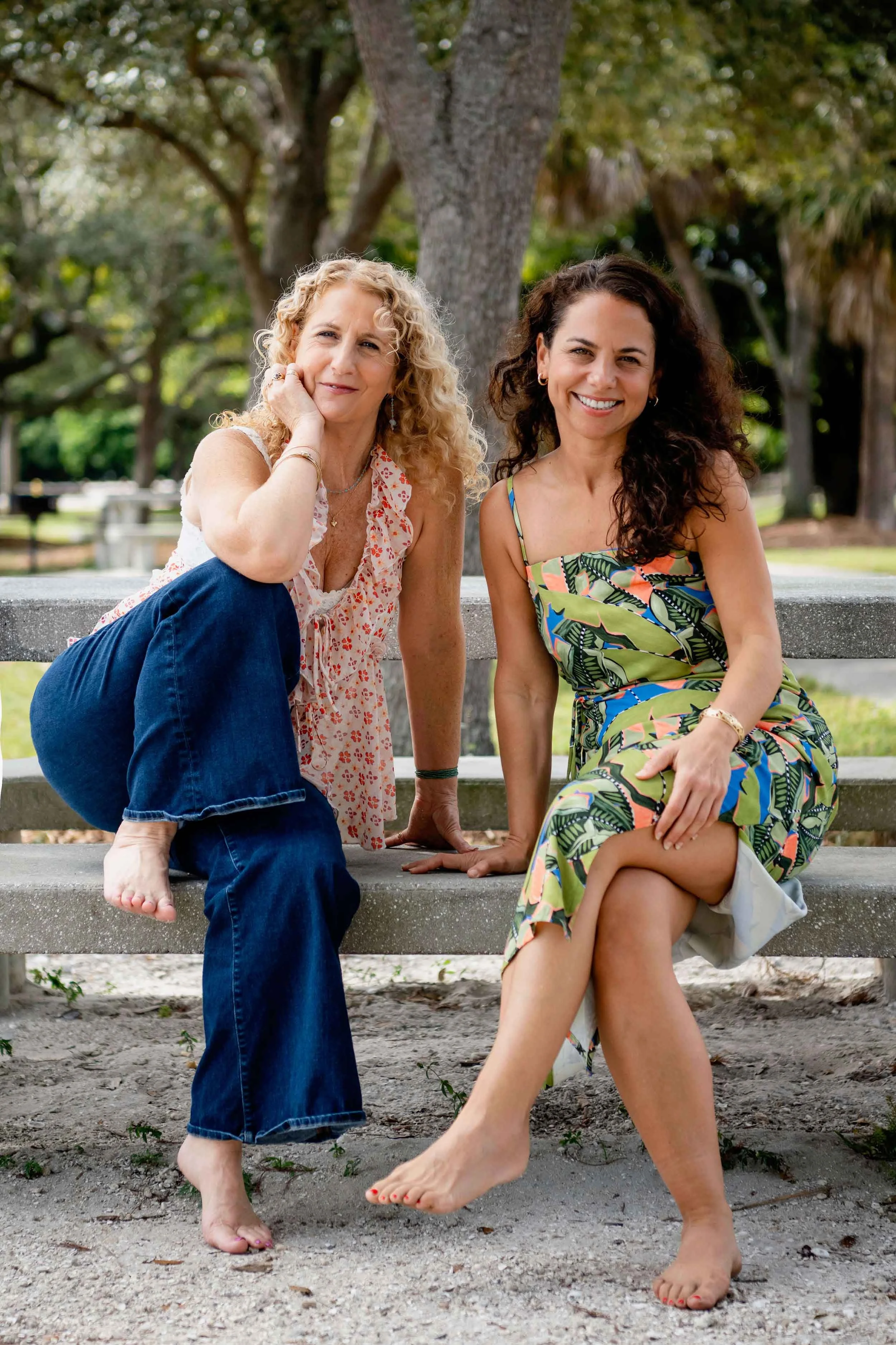 Maureen Delorenzo & Jozi Radus sitting on a park bench outdoors, surrounded by trees, smiling at the camera.