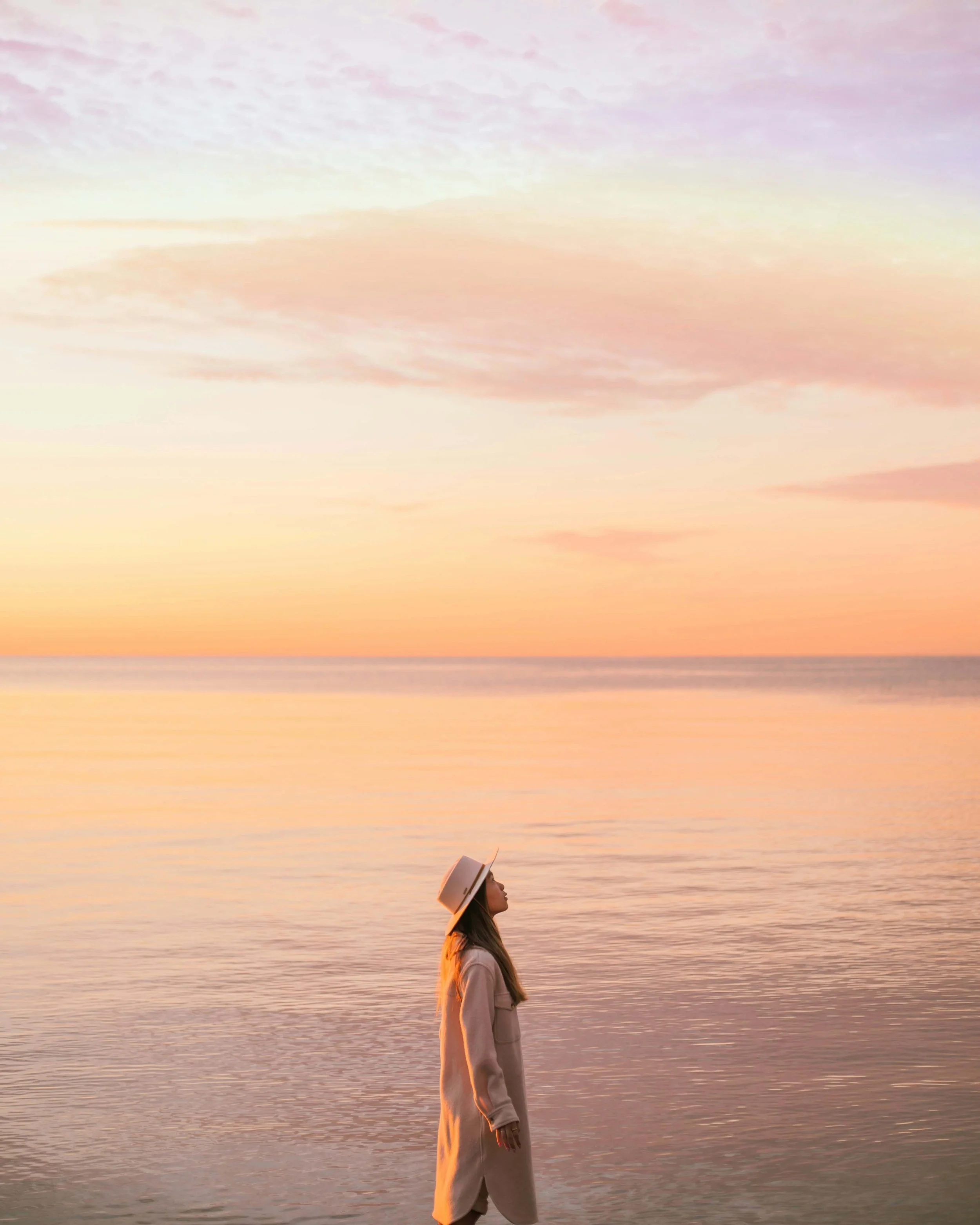 A woman in a coat and hat standing at the edge of calm water during sunset, with pastel-colored sky and clouds in the background.
