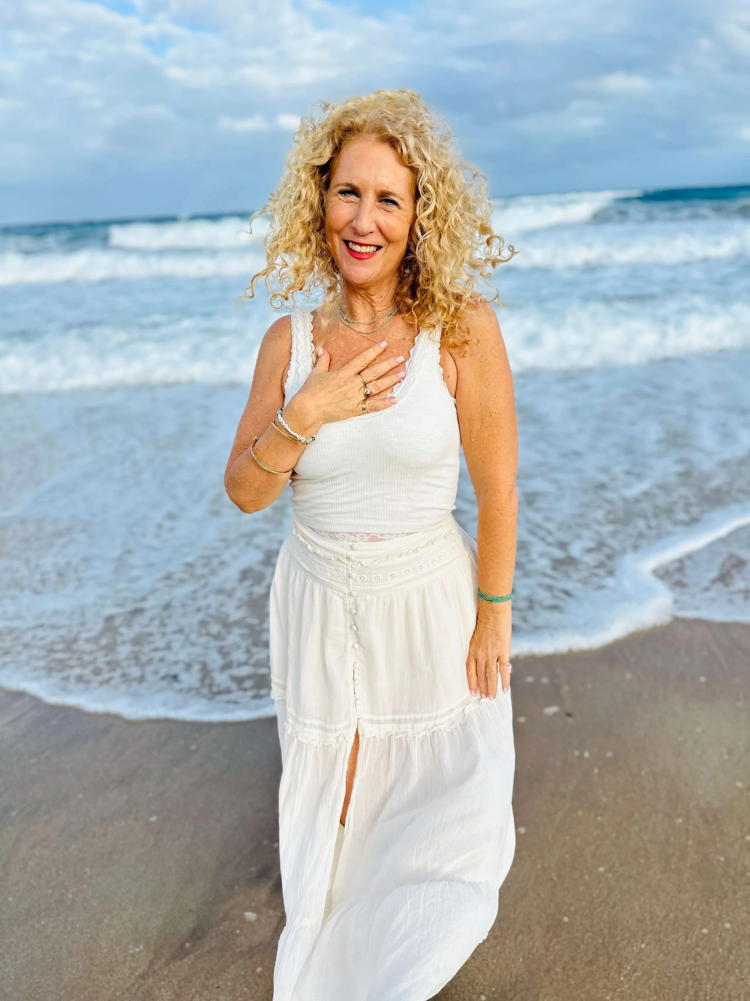 Jozi Radus standing on a beach near the ocean, wearing a white sleeveless top and a long white skirt, smiling and touching her chest.