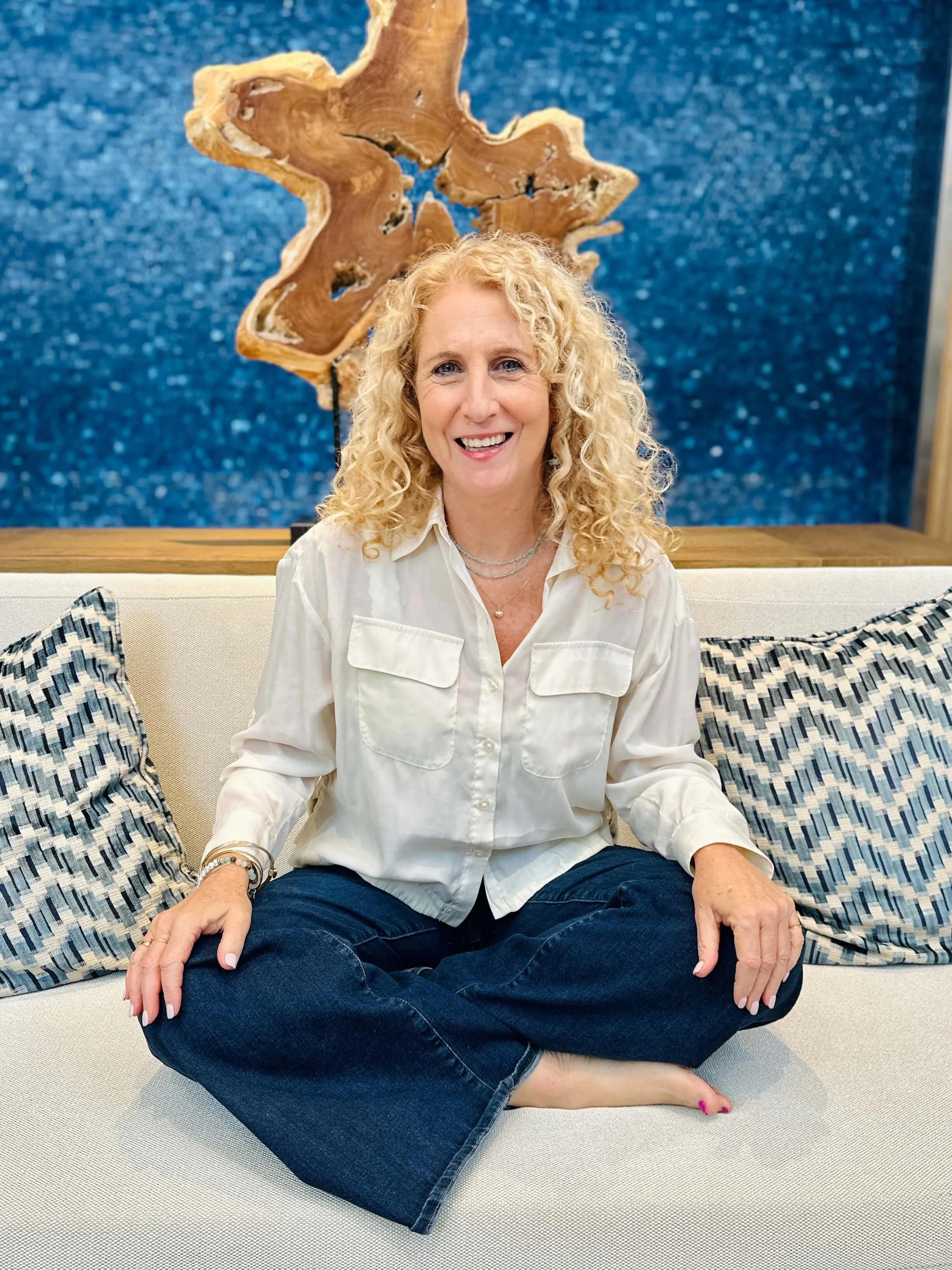 Jozi Radus sitting cross-legged on a cream-colored couch, wearing a white button-up shirt and dark jeans, with patterned pillows behind her and a wooden art piece on a blue textured wall in the background.