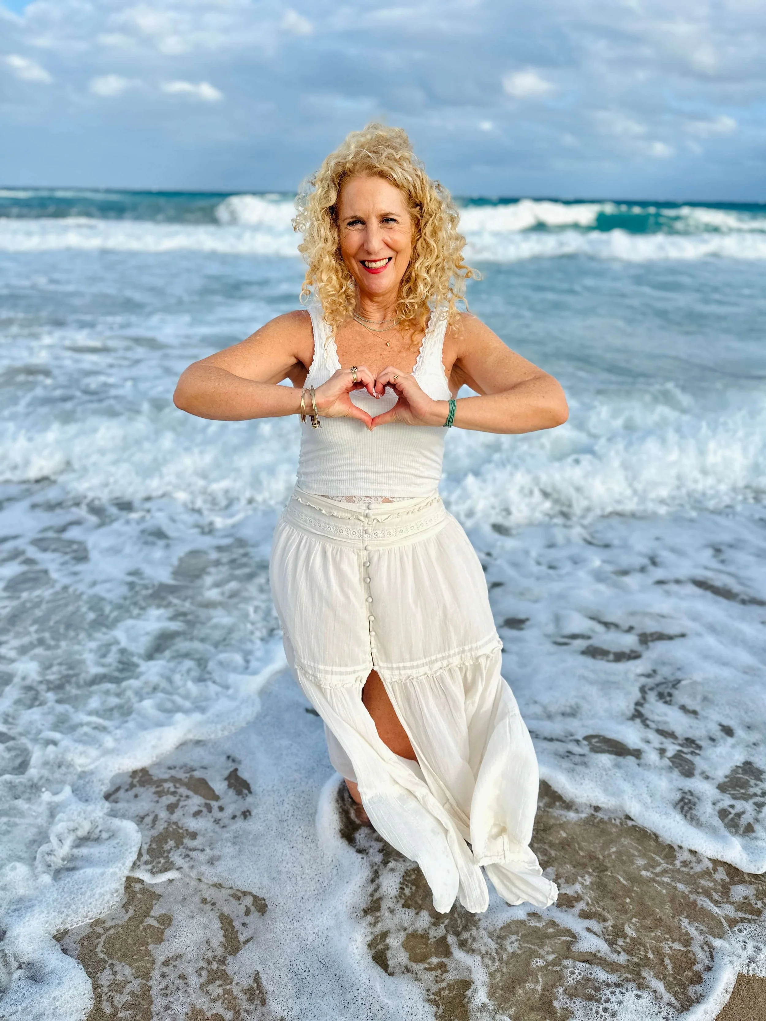Jozi Radus standing in the ocean at the beach, forming a heart shape with her hands. She's wearing a white sleeveless top and a long white skirt with a slit. Waves are crashing around her, and the sky is partly cloudy.
