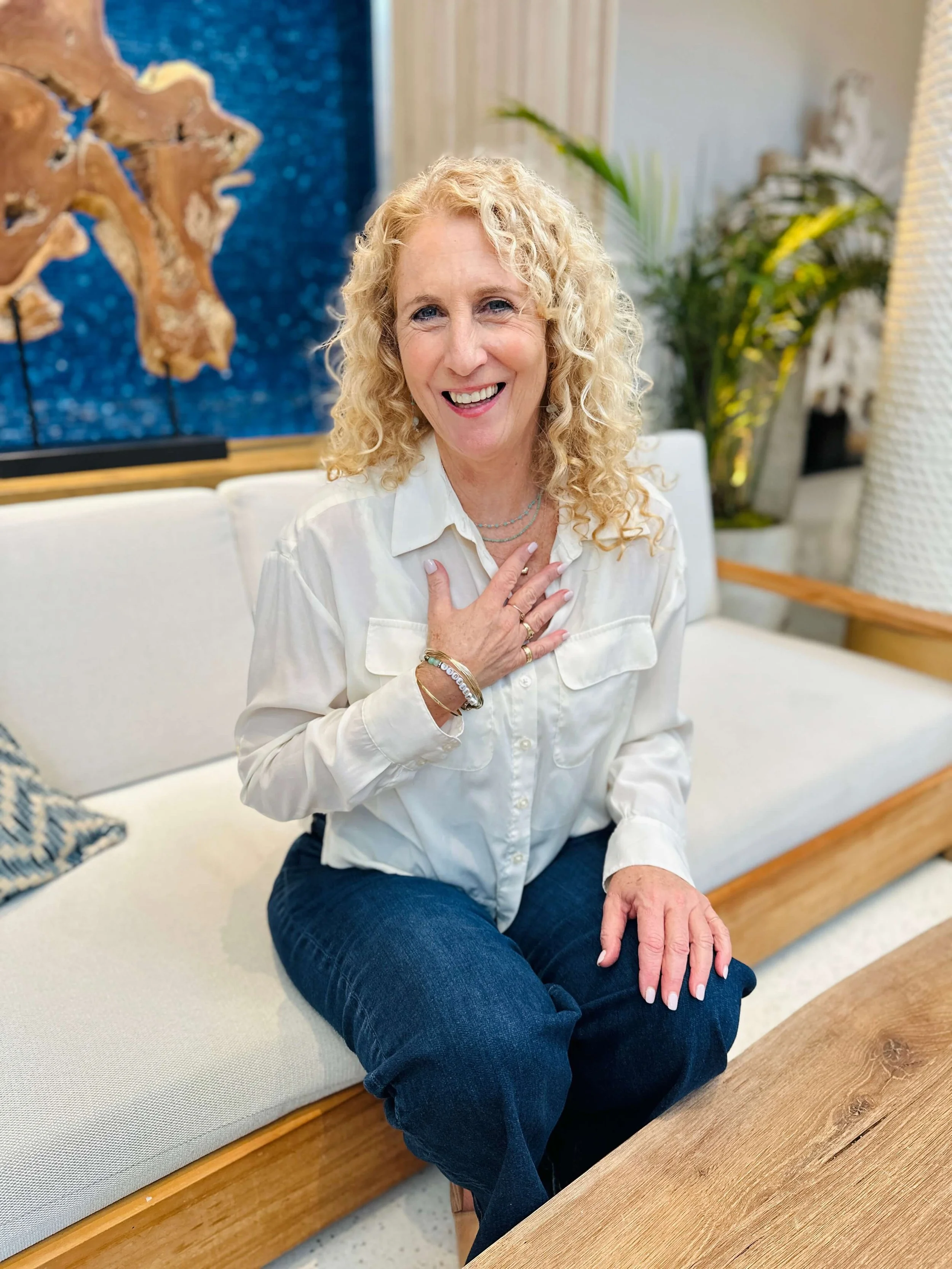 Jozi Radus wearing a white shirt and dark jeans, sits on a white couch with a light wood frame, smiling.