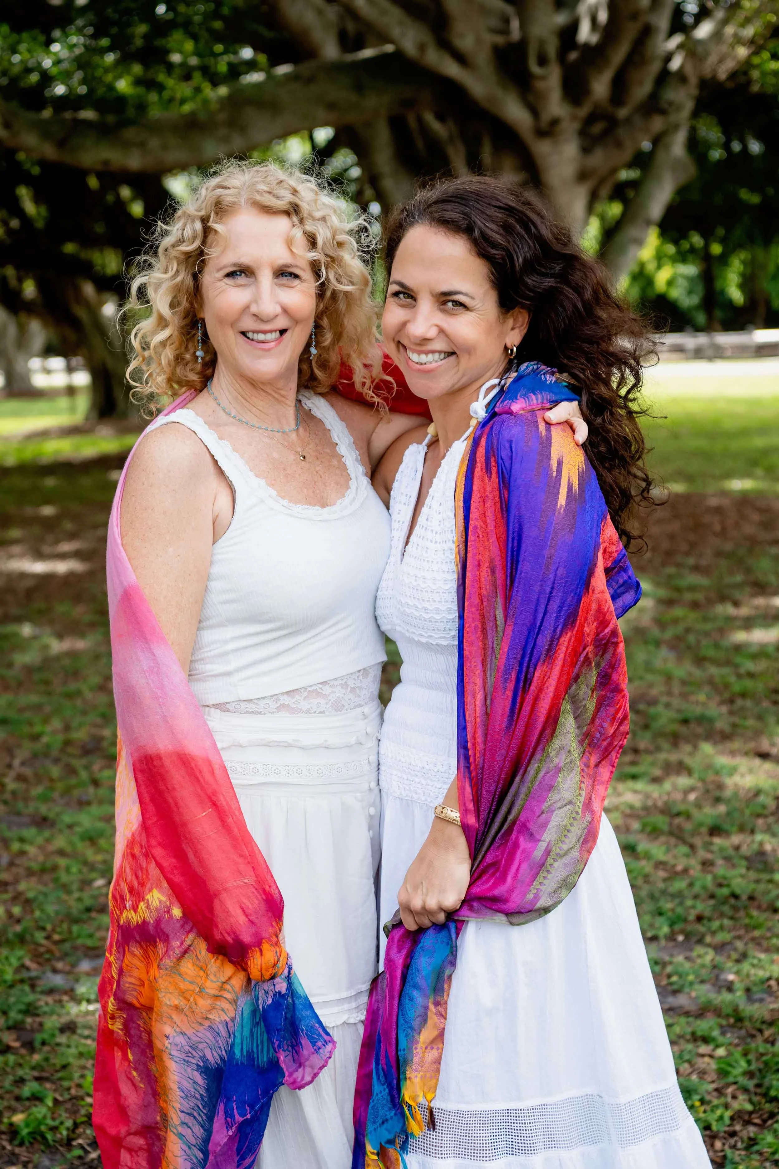 Maureen Delorenzo & Jozi Radus smiling outdoors, standing in front of a large tree with lush greenery, both wearing white dresses and colorful shawls.