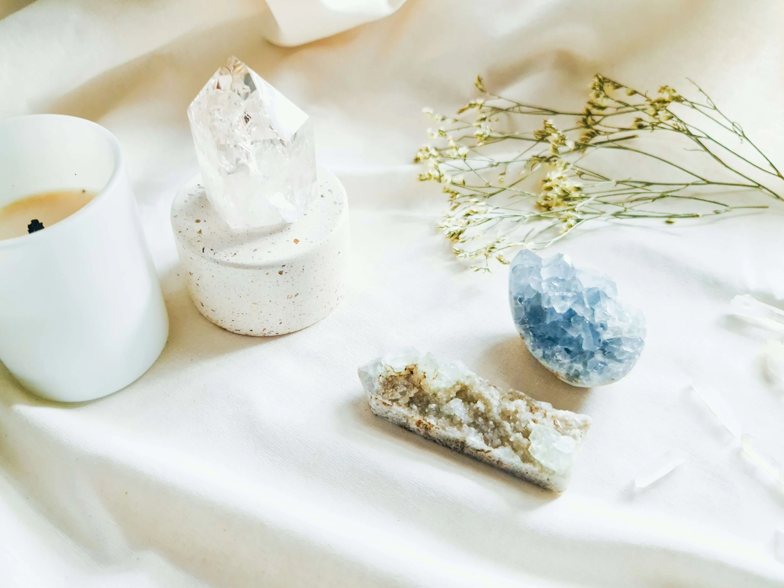 A collection of crystals, a white candle, and dried flowers arranged on a light-colored fabric surface.