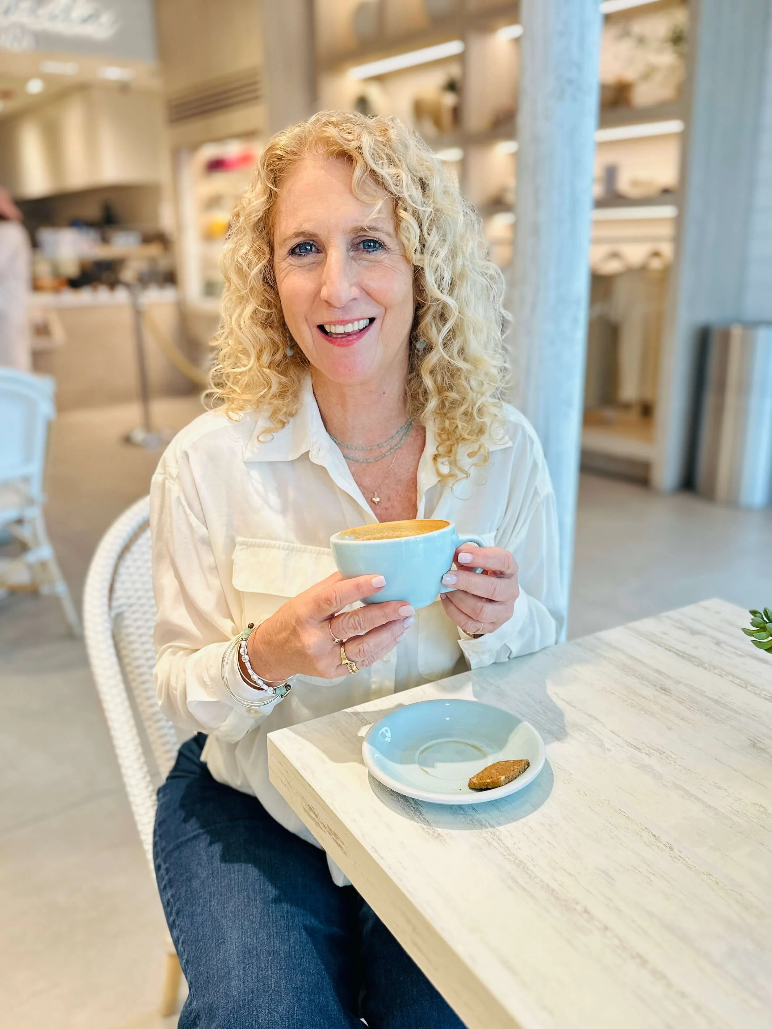 Jozi radus wearing a cream-colored blouse and jewelry, sitting at a light-colored table with a cup of coffee and a cookie in a cafe.