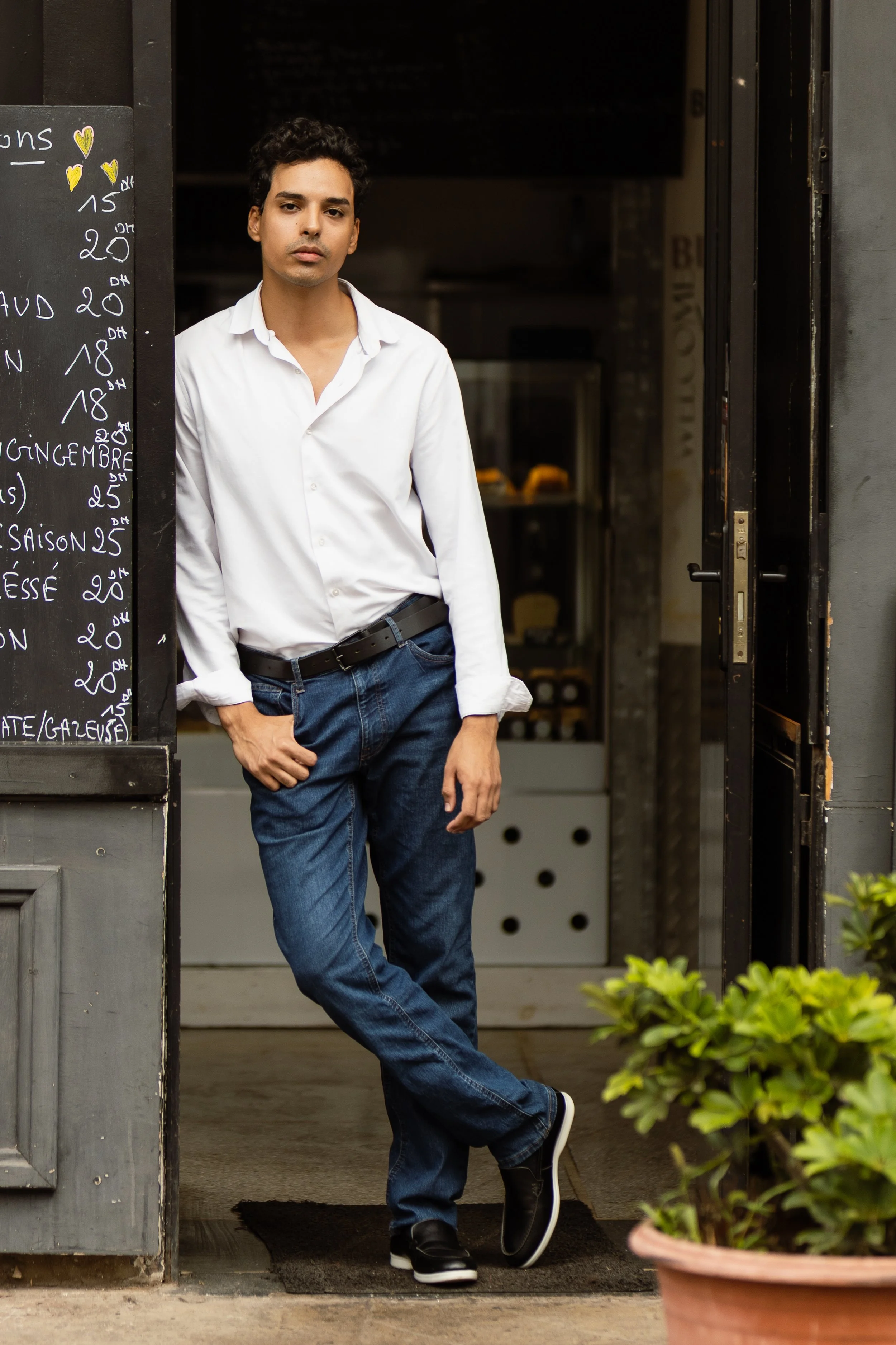 A young man in a white shirt and jeans leaning against a doorway outside a cafe or restaurant.