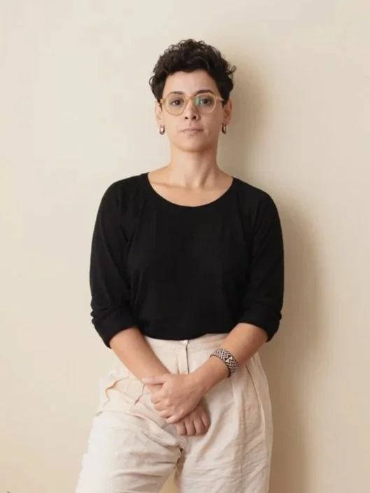 Mulher com cabelo curto e cacheado, óculos, vestindo uma blusa preta de manga longa e calça bege, de pé contra uma parede clara.