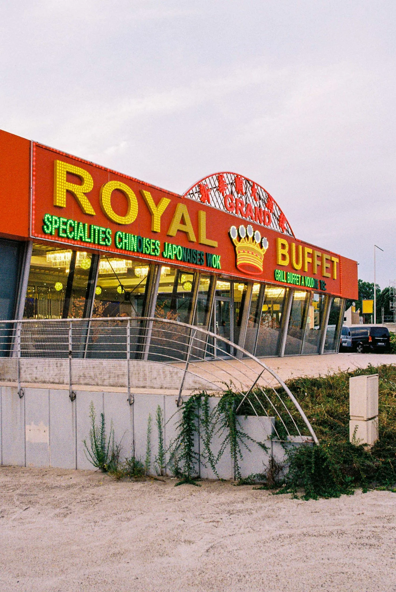 Retro-style building with a large orange and yellow sign reading 'ROYAL BUFFET' and an illuminated neon marquee, featuring a crown graphic, advertising Japanese and Chinese cuisine, sushi, and wok dishes, with a parking lot and overgrown plants in th