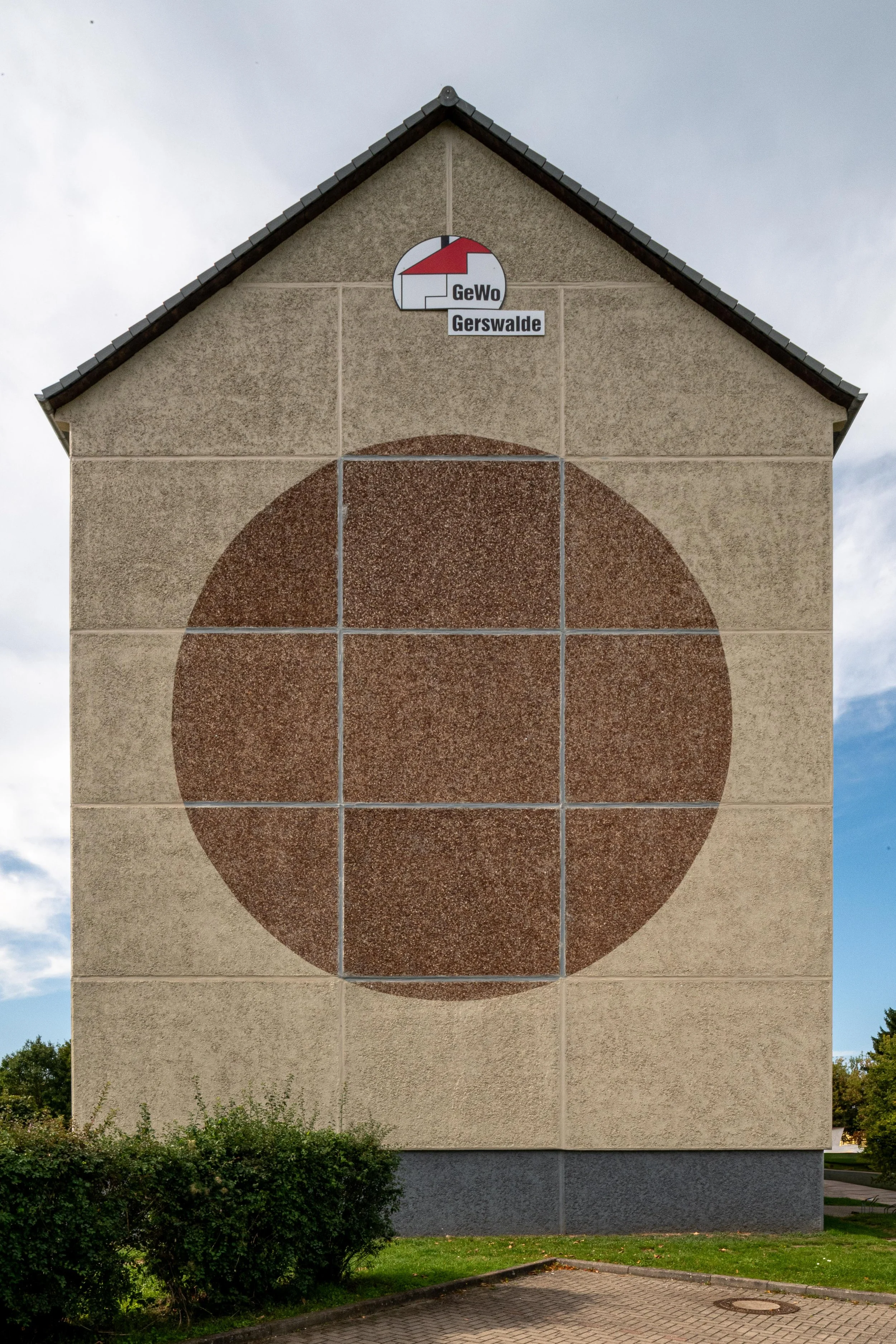 A large, patterned wall on a building featuring a brown circle inside a beige square, with a small gauge at the top and the words 'GeWo Gerswalde' beneath it.