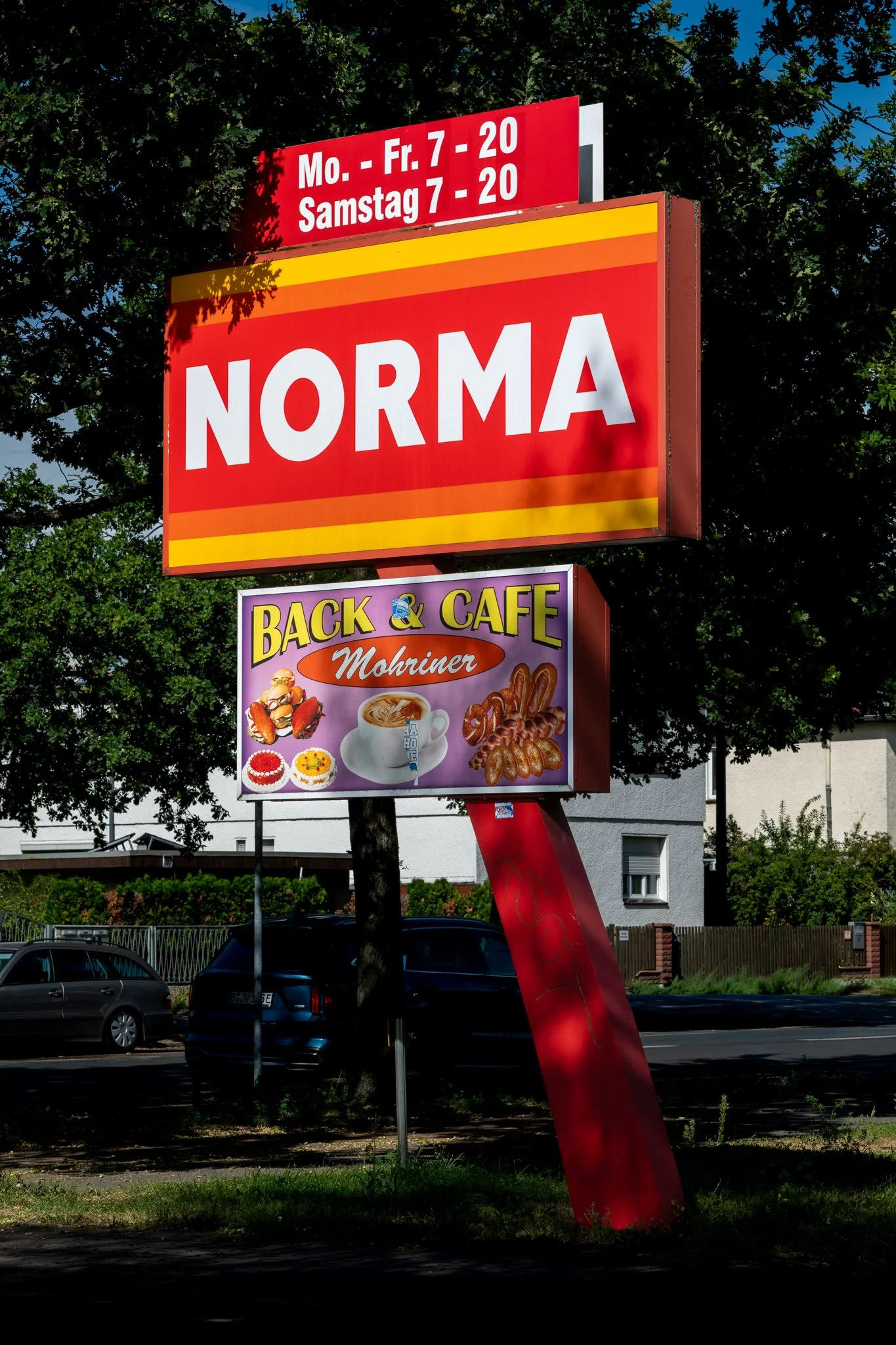 A roadside sign with a red background displaying business hours in German, the word 'NORMA' in large white letters, and a sign for a cafe with images of food and beverages on a purple background.