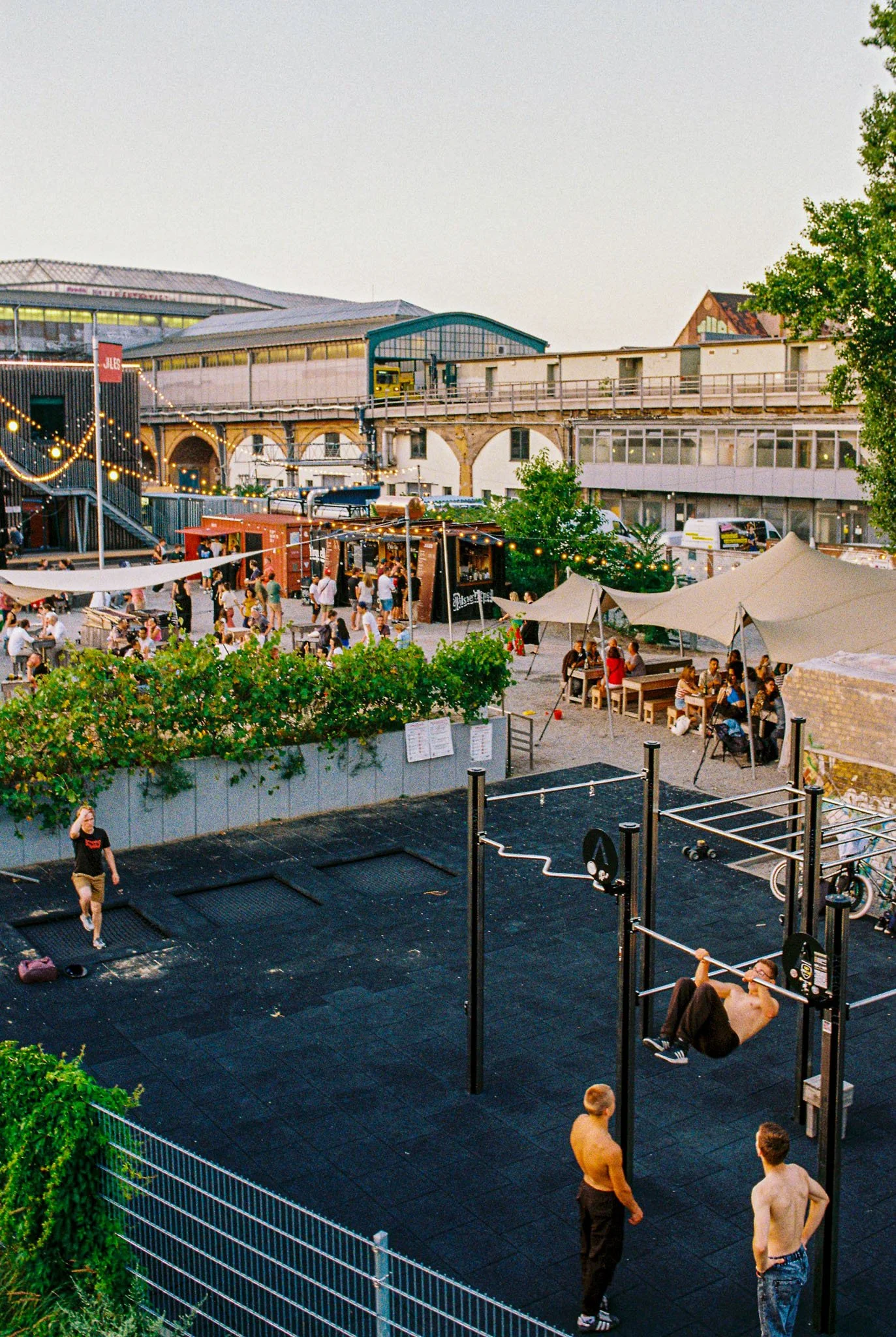 An outdoor recreational area with people walking, sitting under umbrellas, and a man exercising on a pull-up bar. In the background, there is a market or festival with strings of lights, food stalls, and a high-rise structure with an elevated railway