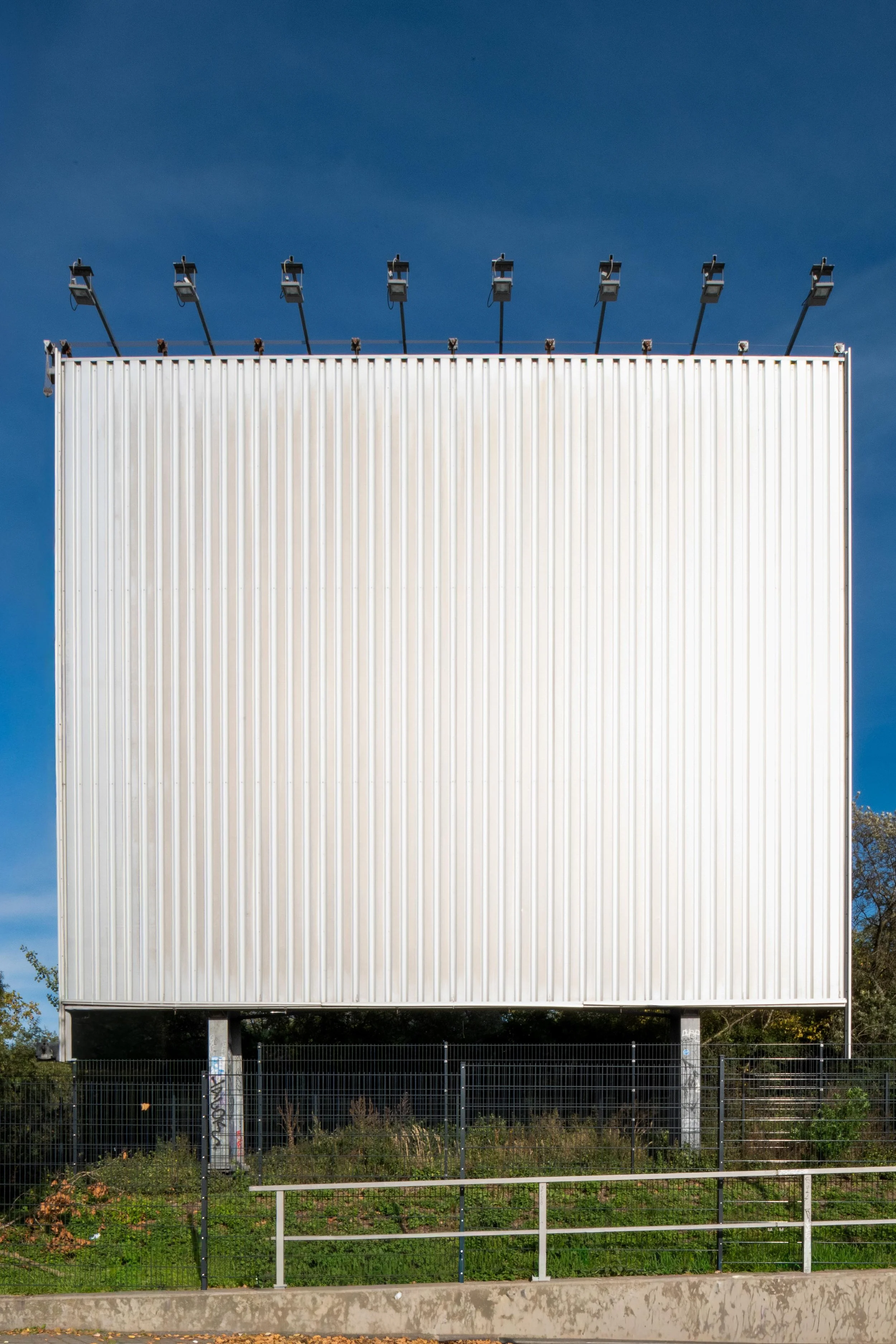 A large, white blank billboard with several lights mounted on top, situated outdoors against a blue sky. There is a fence and some greenery at the base.