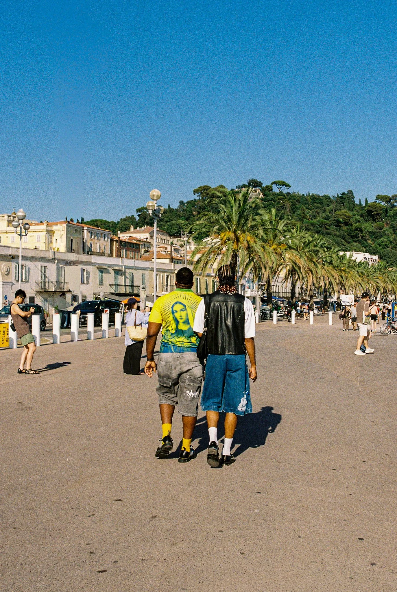 Two men walk hand in hand along a sunny promenade with palm trees, other pedestrians, parked cars, and a hillside with houses in the background.