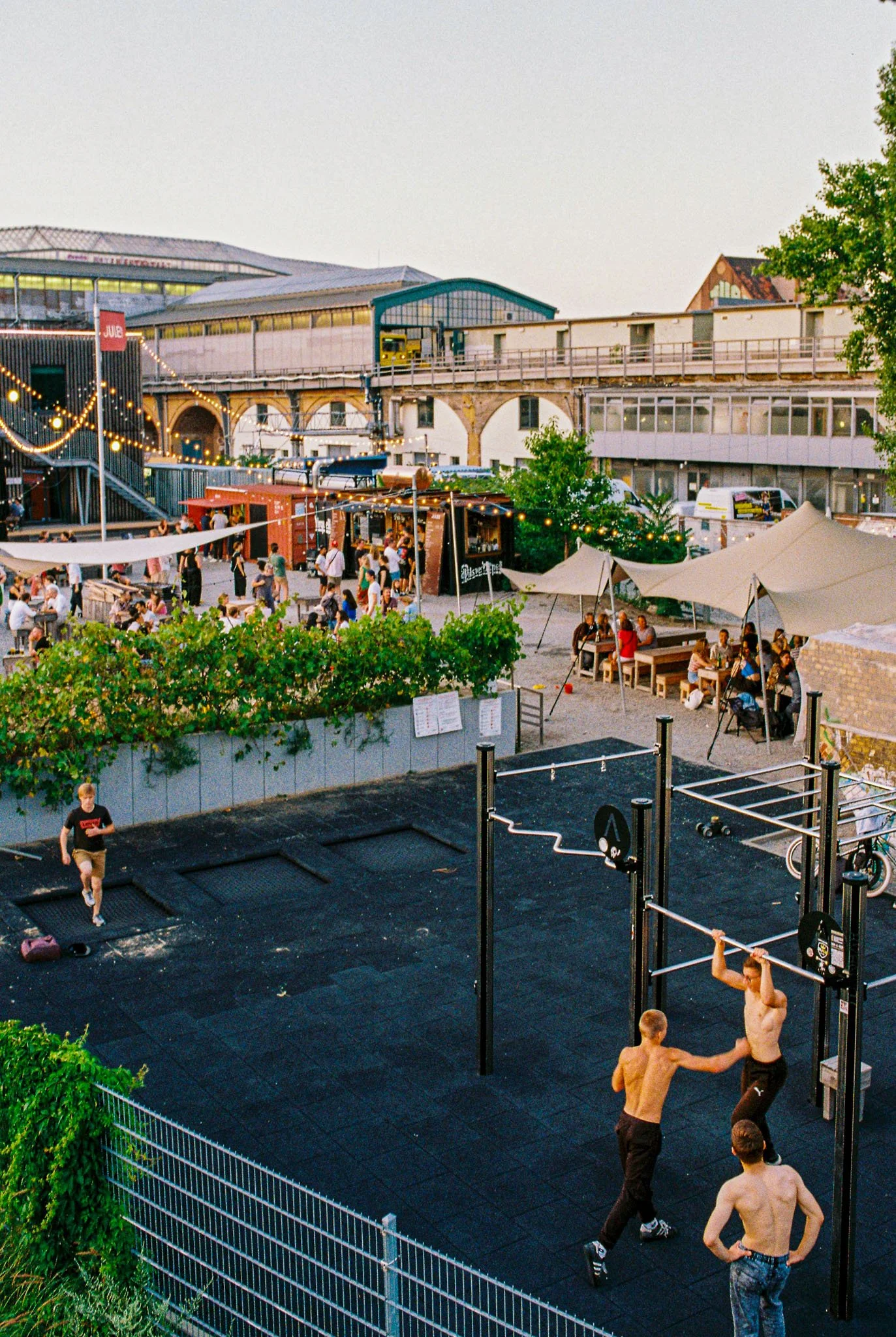 Outdoor social area with string lights, food stalls, tents, and people sitting and walking. A small playground with two shirtless men lifting weights and a boy jumping on a trampoline in the foreground.