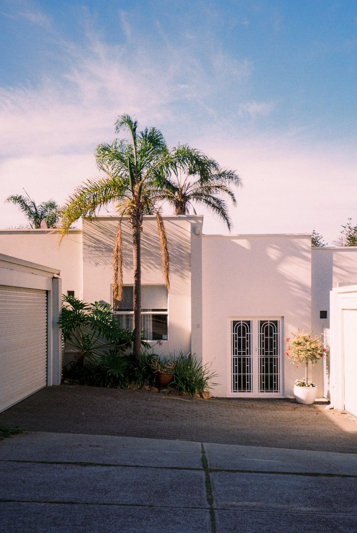 A modern white house with palm trees in front, a small potted tree on the right side, and a partly open garage door on the left. The sky is clear with some clouds.