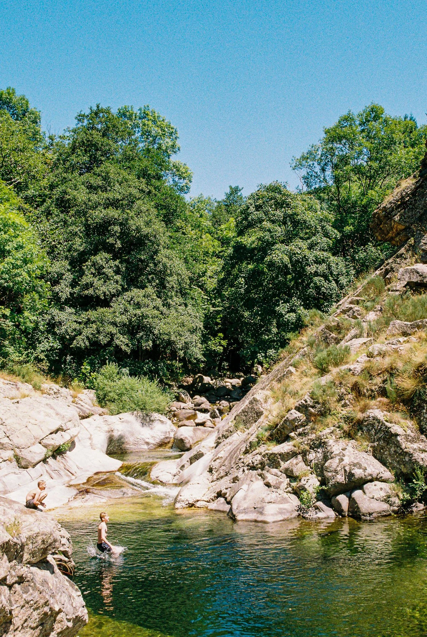 Two children playing in a clear river surrounded by large rocks and green trees under a bright blue sky.