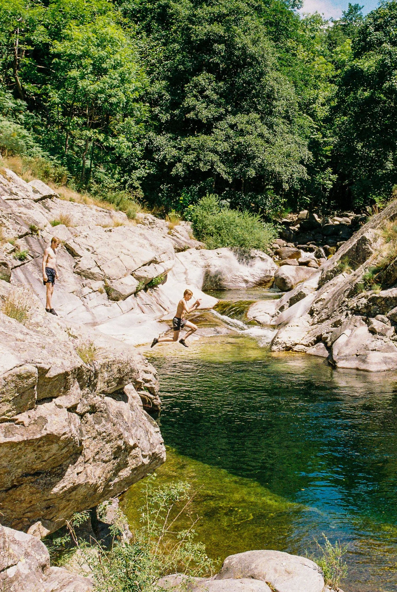 Two boys jumping into a creek surrounded by large rocks and lush green trees.