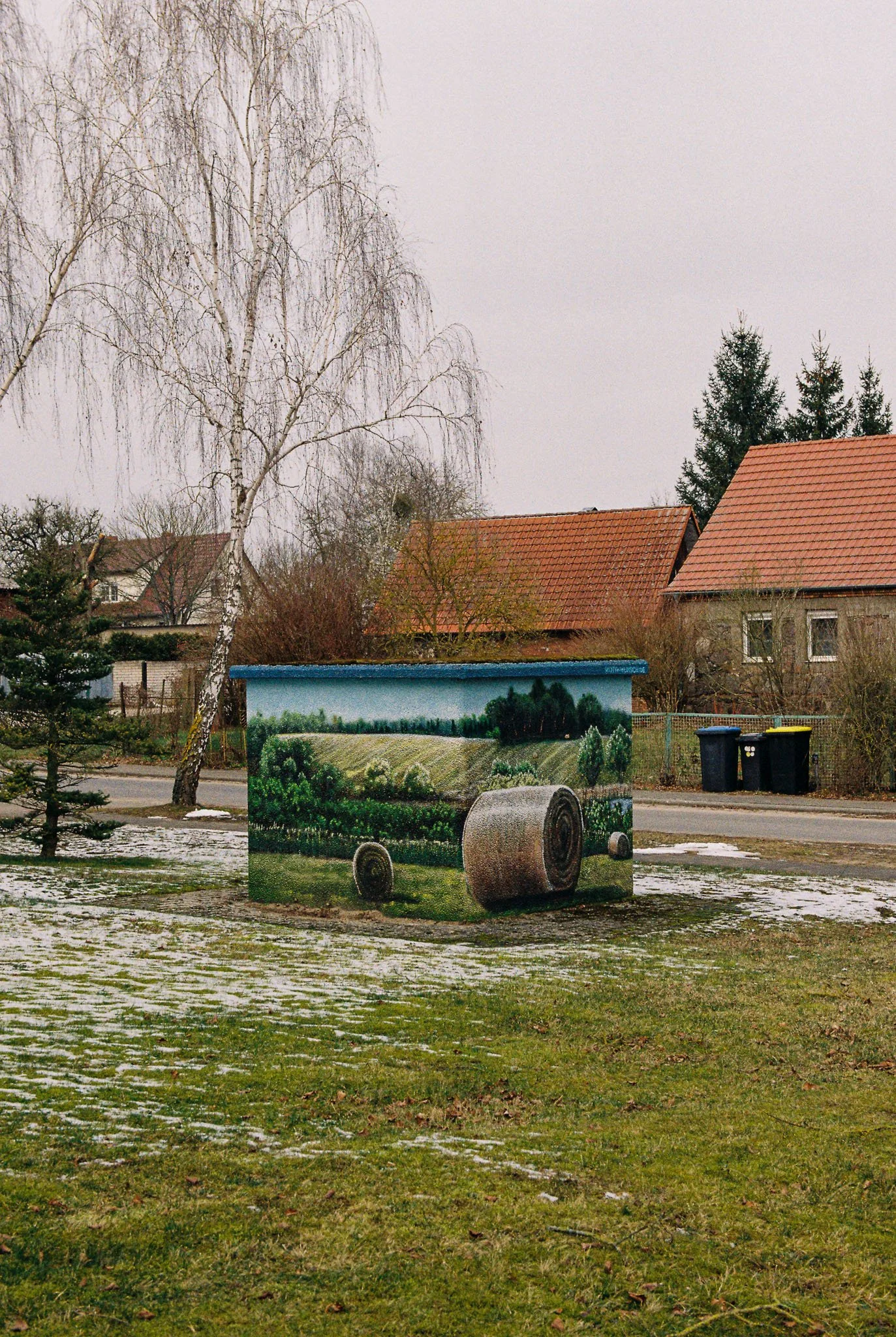 A mural on a small structure in a yard features rolling fields, trees, and sky, with concrete roller sculptures placed in front. The yard has green grass with patches of snow, trees, and neighboring houses with red roofs in the background.