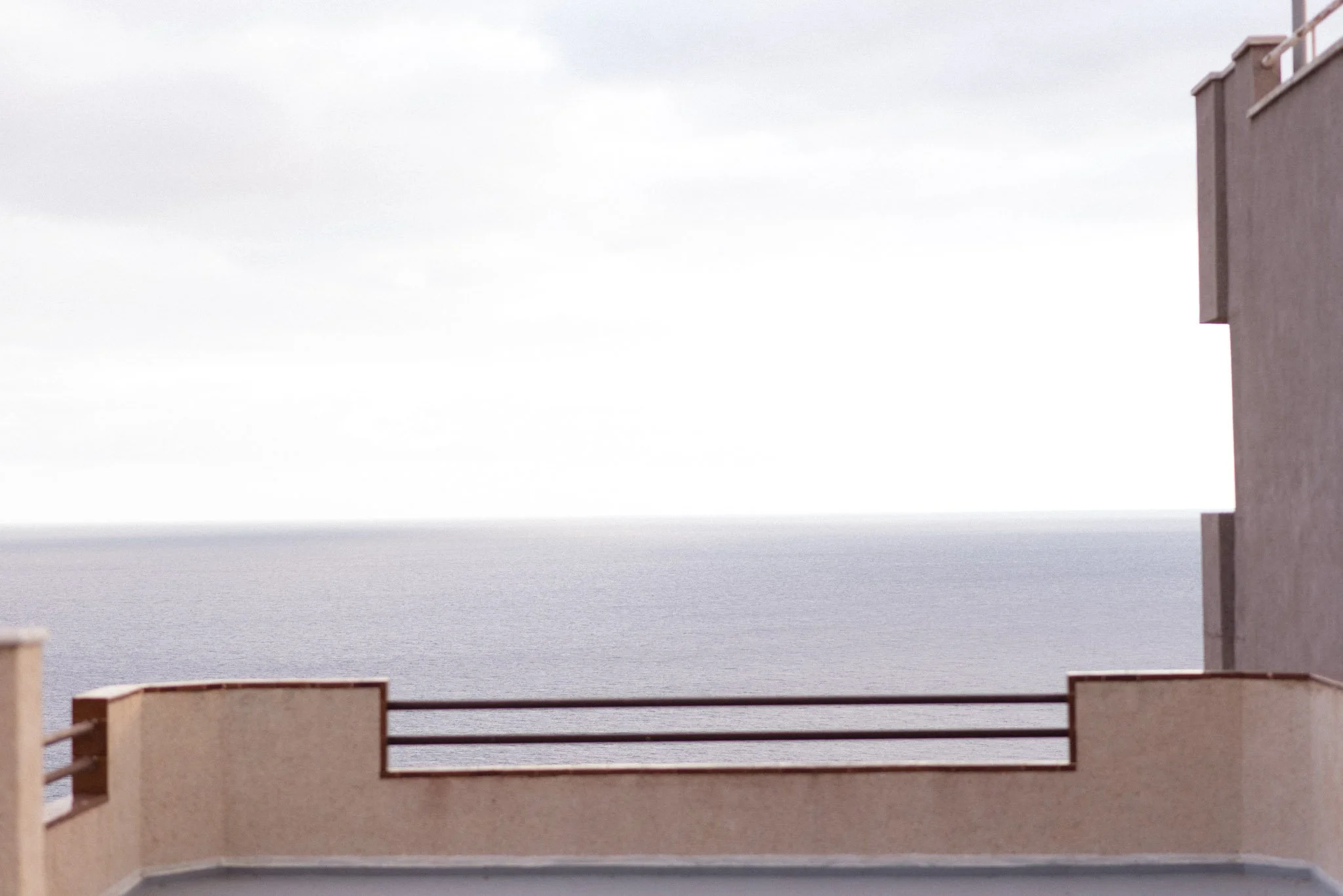 View of an ocean or large body of water seen from a balcony or terrace of a building, with a concrete or stucco railing in the foreground and neighboring buildings on the sides, under a cloudy sky.