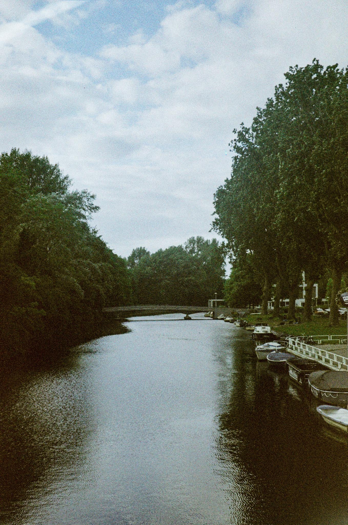 Scenic view of a river with boats docked along the right bank, trees lining both sides, and a cloudy sky overhead.