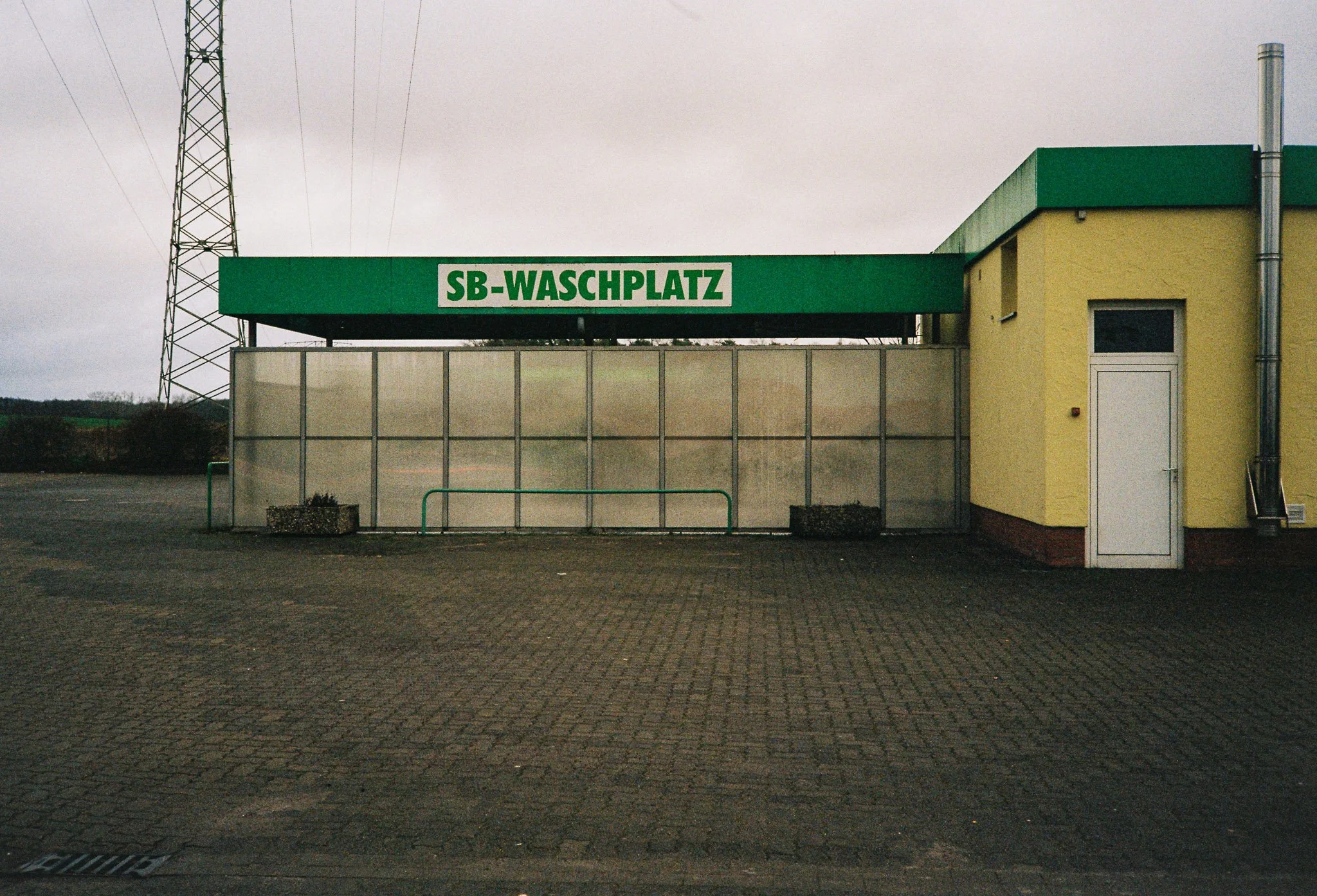 Empty car wash station with a green sign reading 'SB-Waschplatz', located outside a yellow building with a white door, on overcast day.