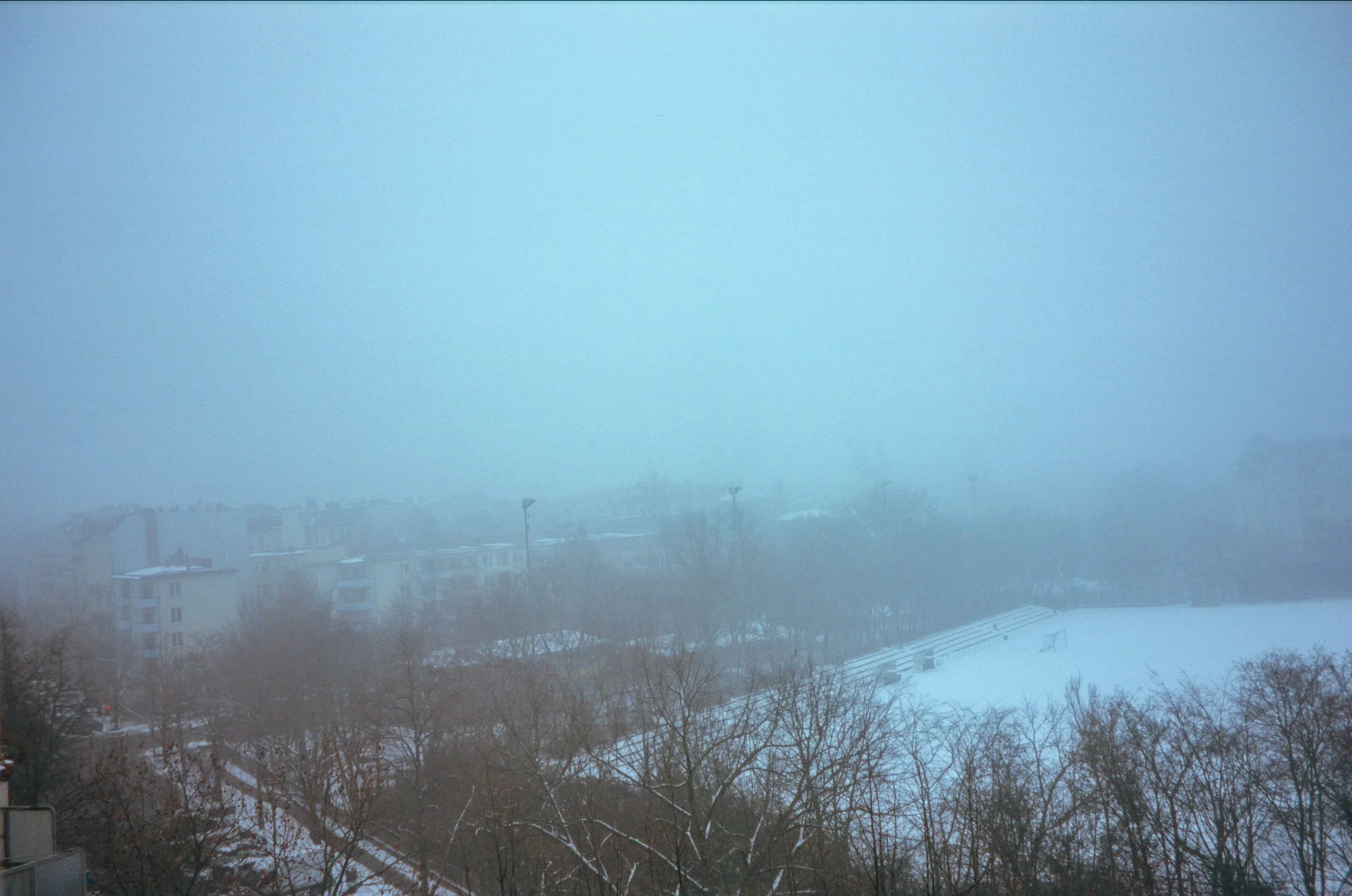 A snowy landscape with an open field, a playground, leafless trees, and a row of apartment buildings in the distance, obscured by fog or heavy snowfall.