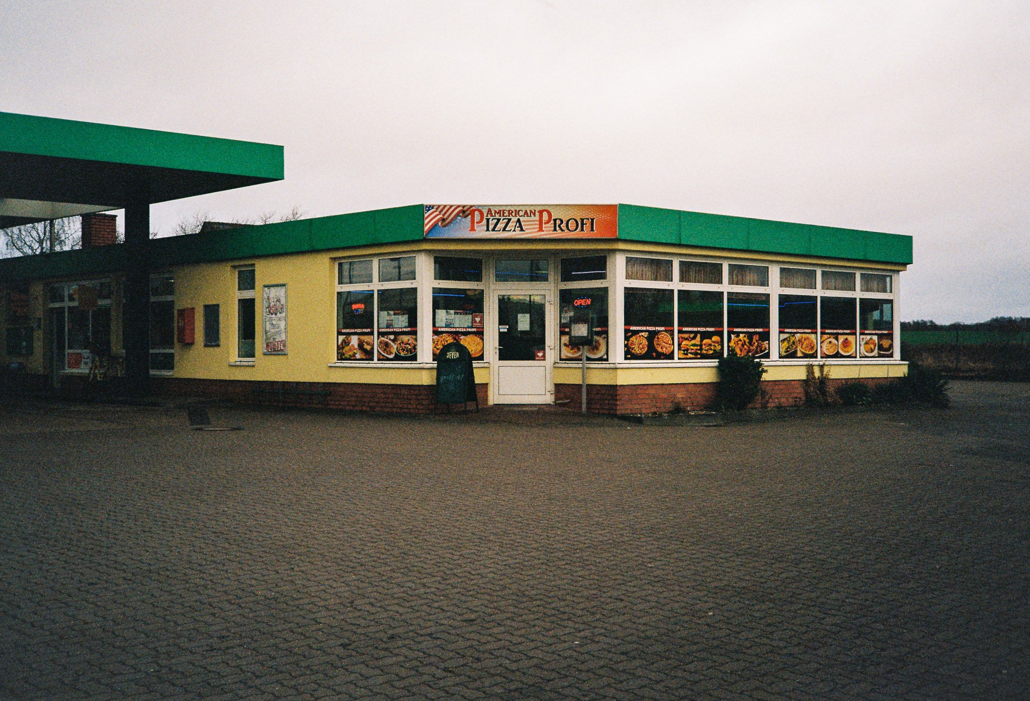 A small restaurant with yellow walls, large windows displaying food pictures, and a green awning. The sign reads "American Pizza Profi".