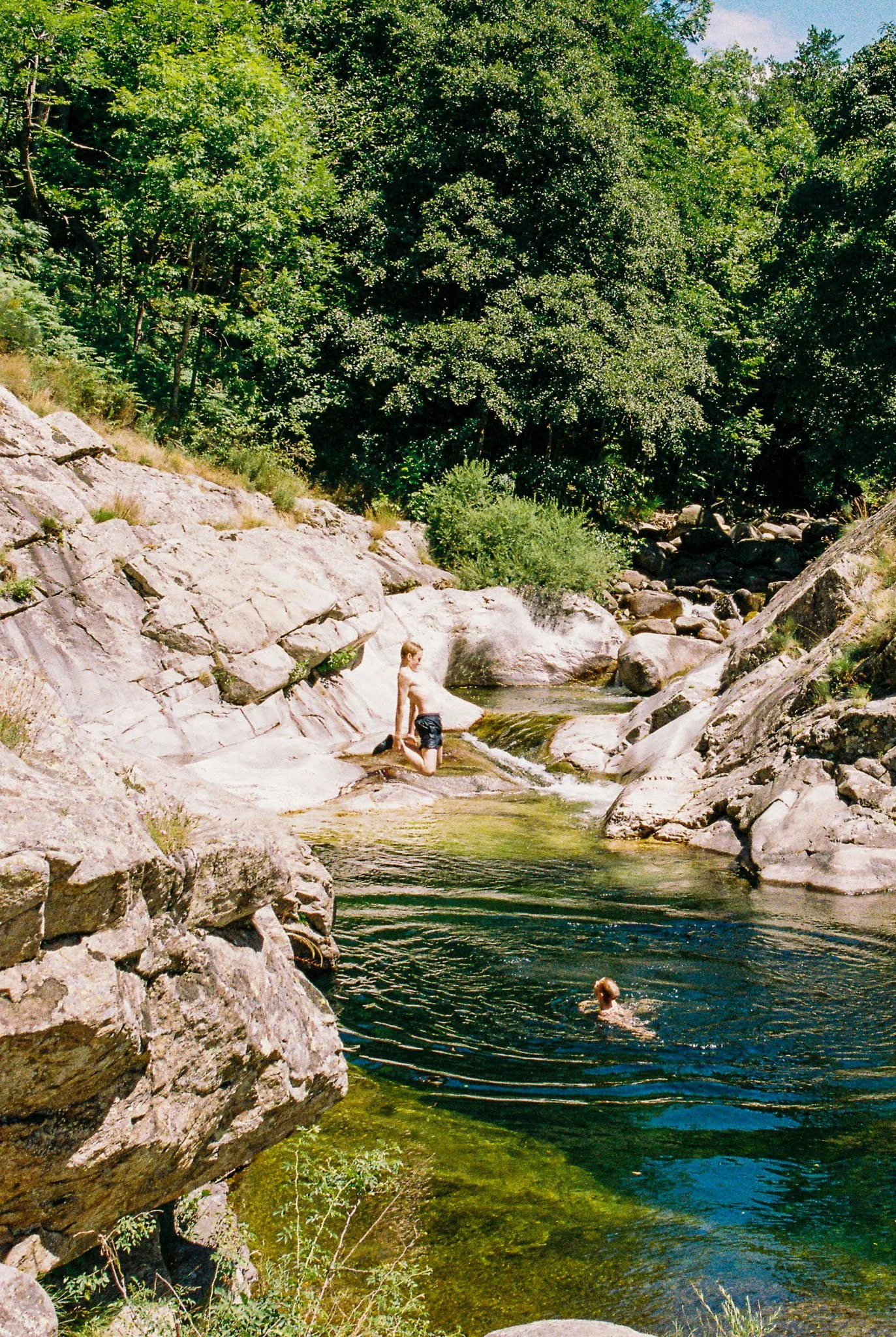 A boy in swim shorts sitting on a rock in a shallow part of a rocky creek, with another person swimming in the water nearby surrounded by trees.