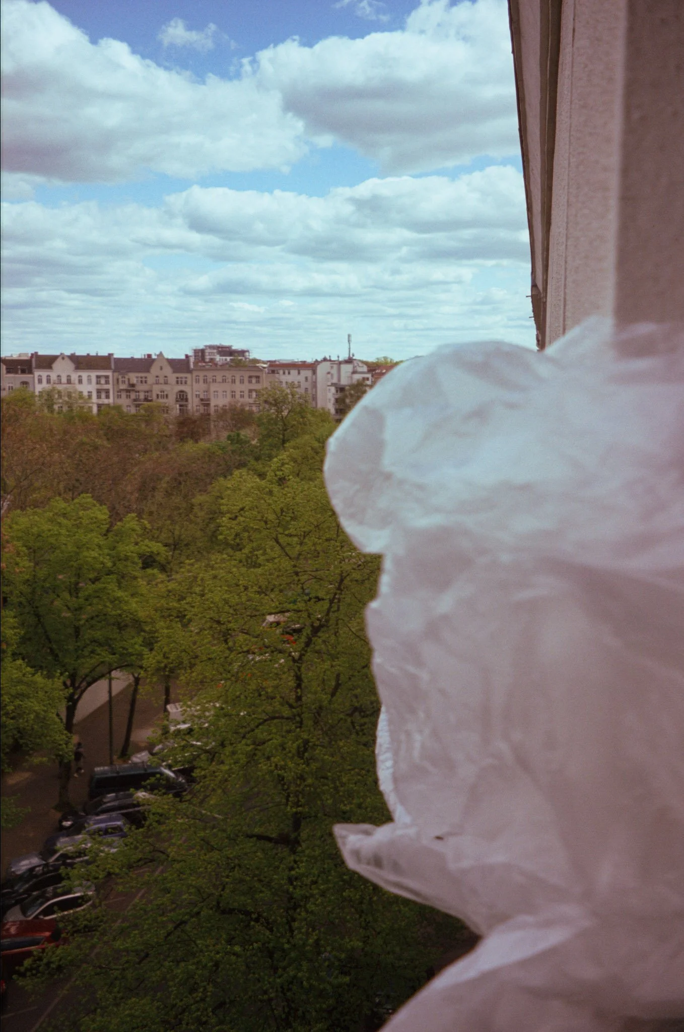 View from a window showing green trees and a parking lot with parked cars, with a cityscape of cream-colored buildings and a partly cloudy sky in the background.