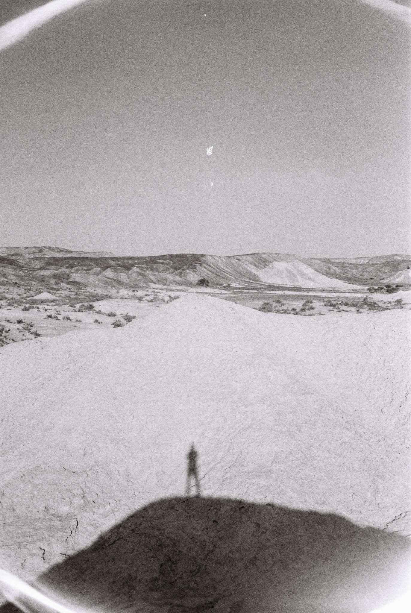 A person standing on a mound of sand or dirt in a desert landscape, with hills in the distance and a clear sky.