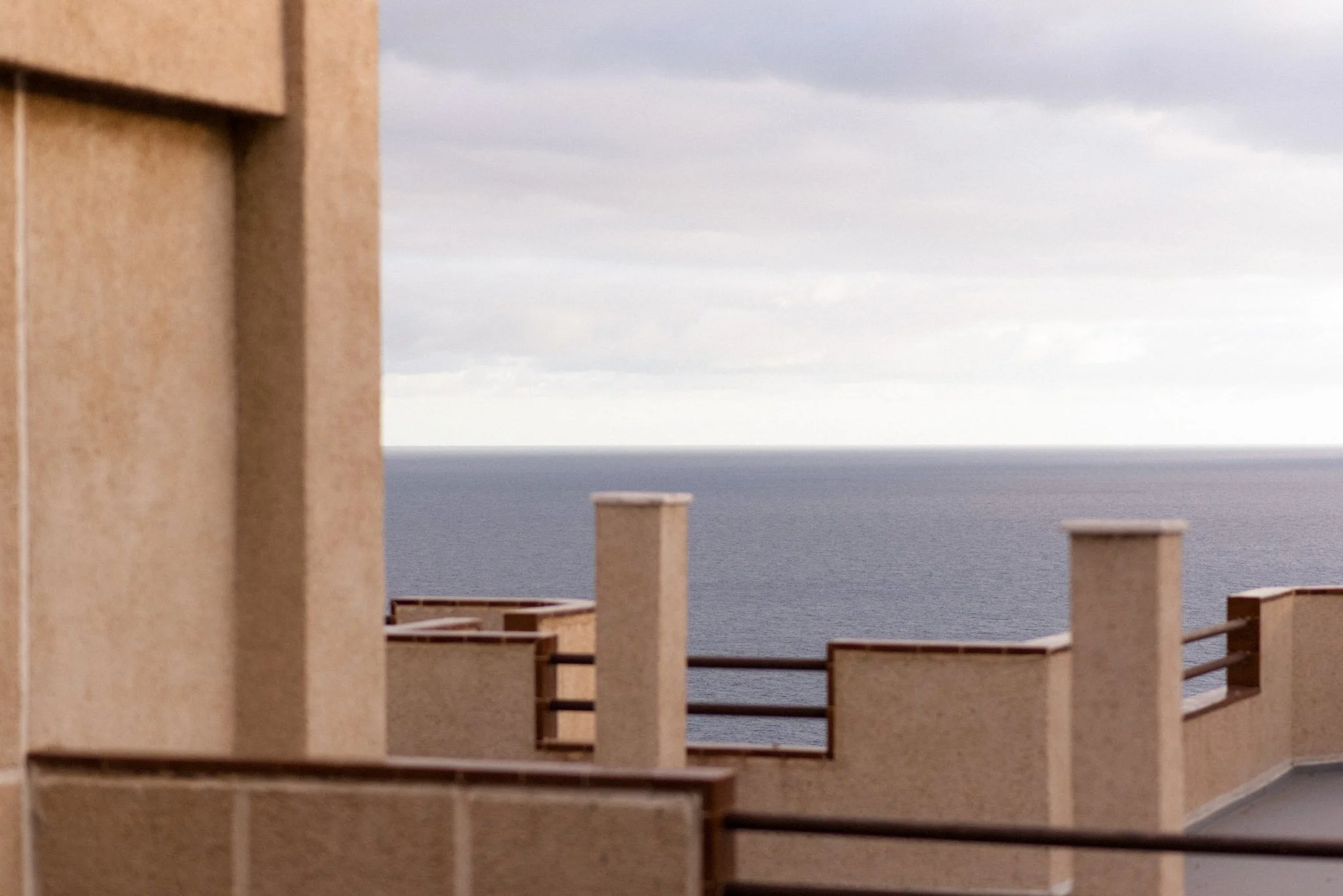 View from a balcony showing beige building with vertical pillars, railing, and a cloudy sky over the ocean in the background.