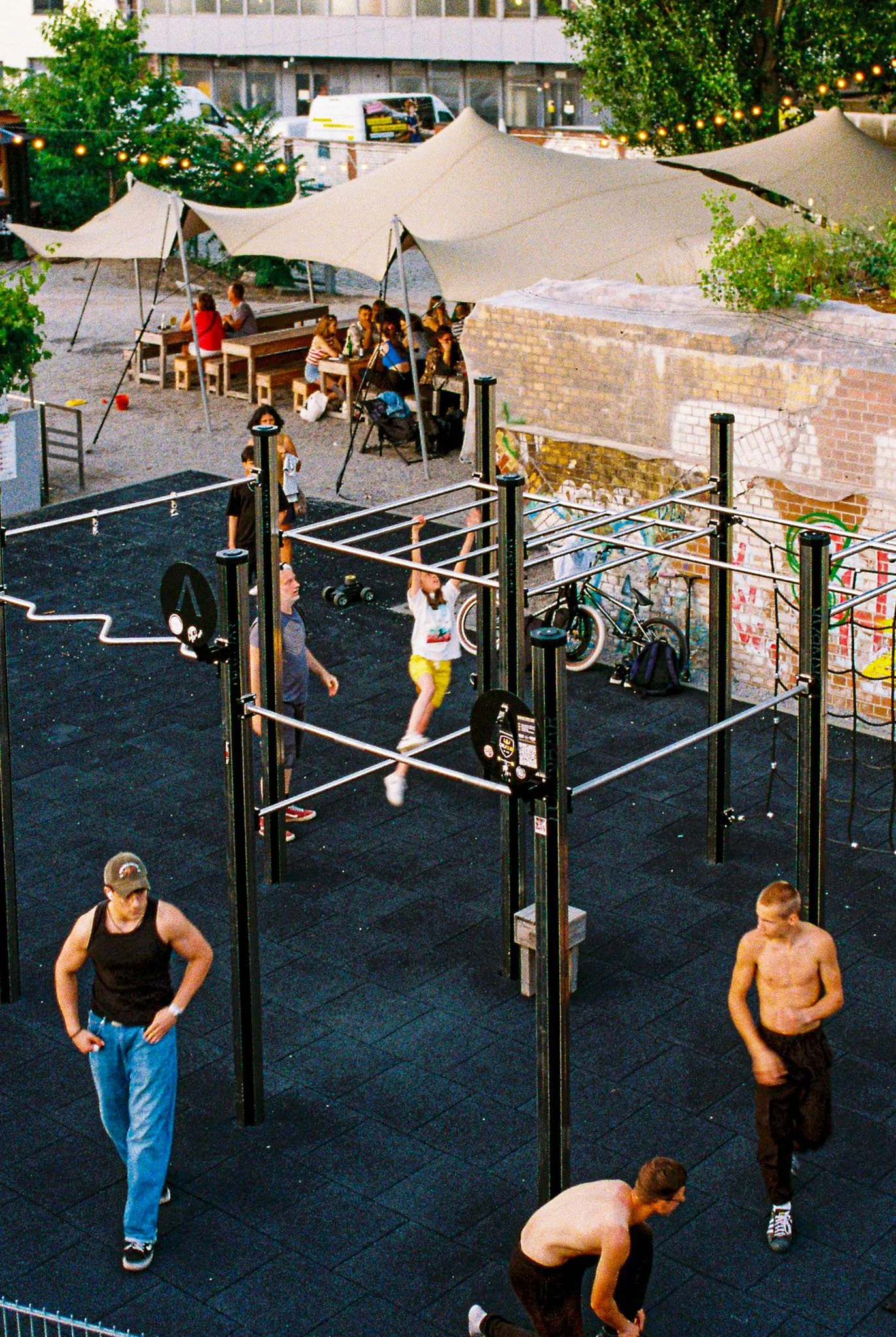People working out and socializing at an outdoor gym and dining area with string lights, umbrellas, and trees.